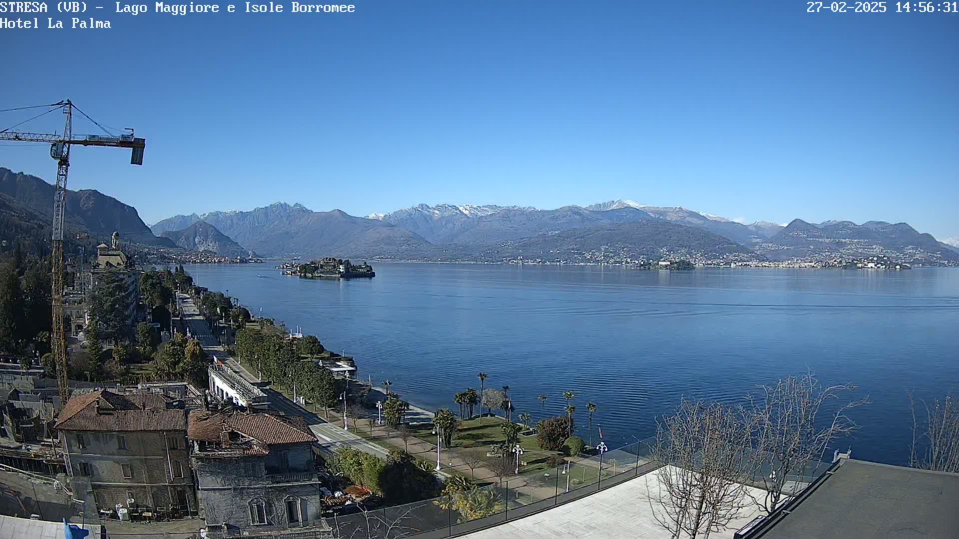 A clear, sunny day overlooks a calm lake with snow-capped mountains in the distance, a small island in the lake, and a shoreline with buildings and vegetation.