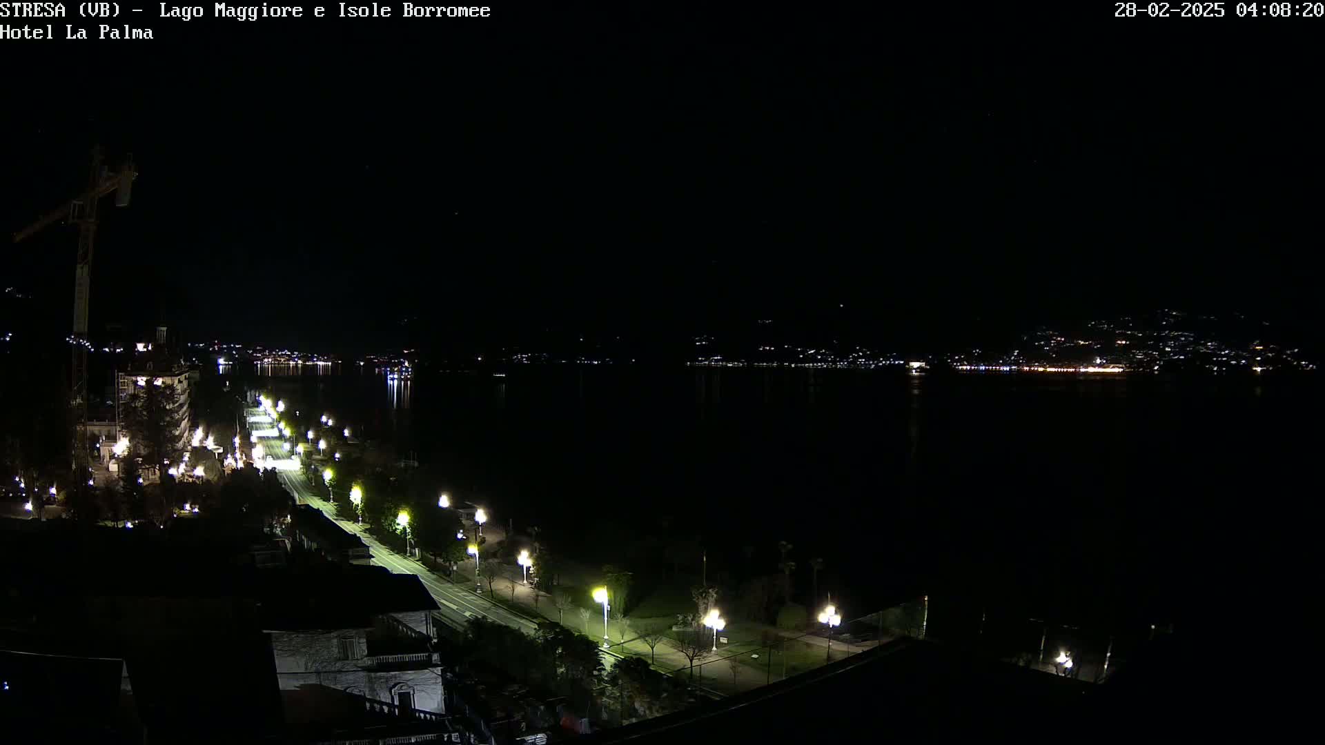 A nighttime view of a lakeside road lined with lights, leading to a dark body of water with faintly illuminated buildings on the far shore.
