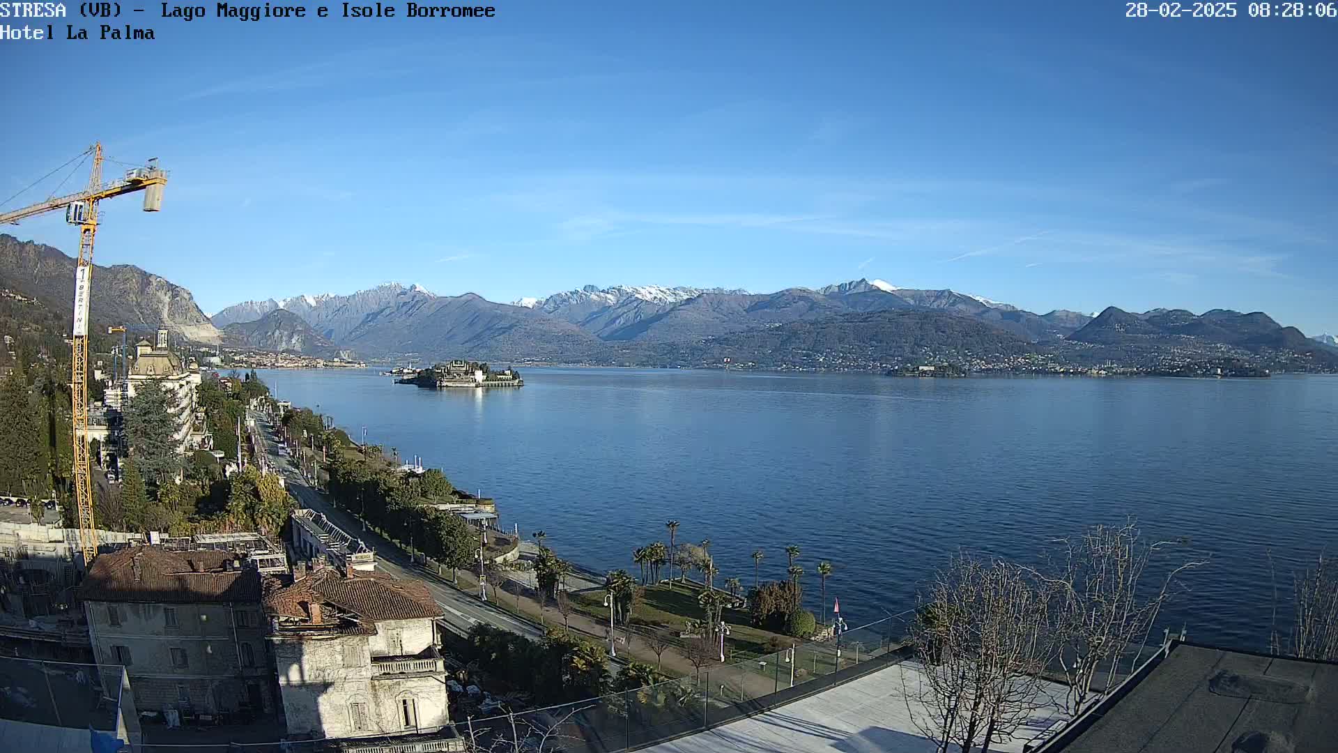 A clear, sunny day overlooking a large lake with snow-capped mountains in the background, a small island in the middle of the lake, and buildings along the lakeshore.