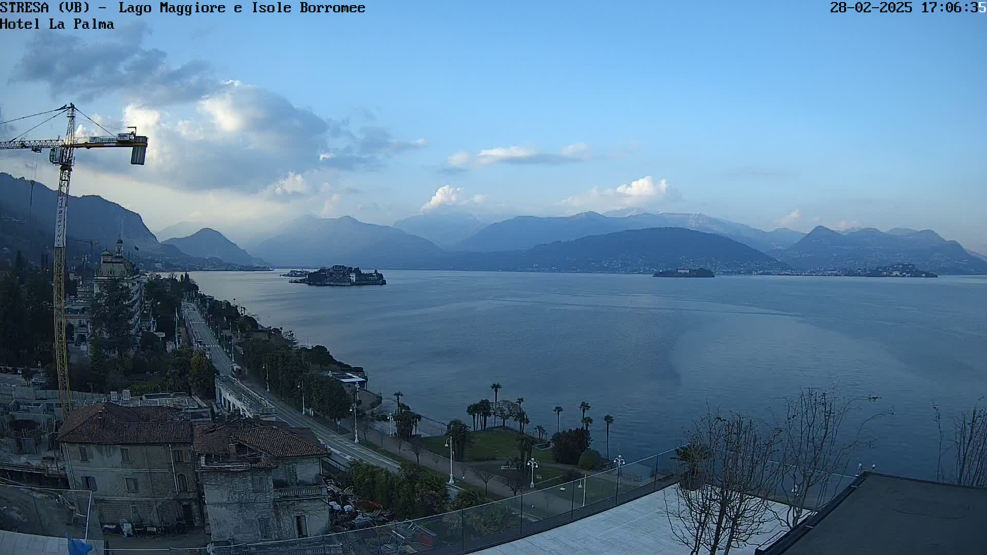 A partly cloudy day overlooks a calm lake with islands and mountains in the distance, a town and construction crane visible in the foreground.