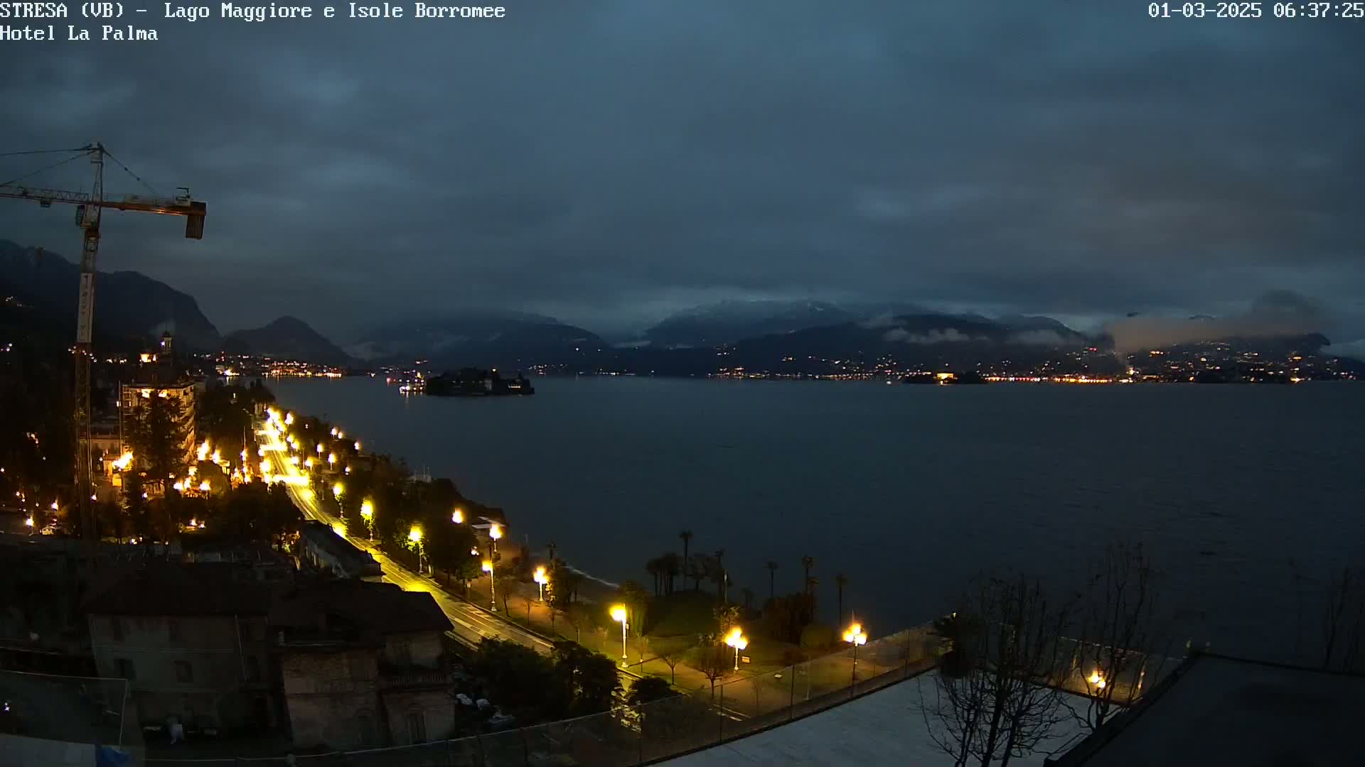A nighttime view of a city's lakeside illuminated streets and buildings, under a dark, cloudy sky, with snow-capped mountains visible in the distance across a calm lake.