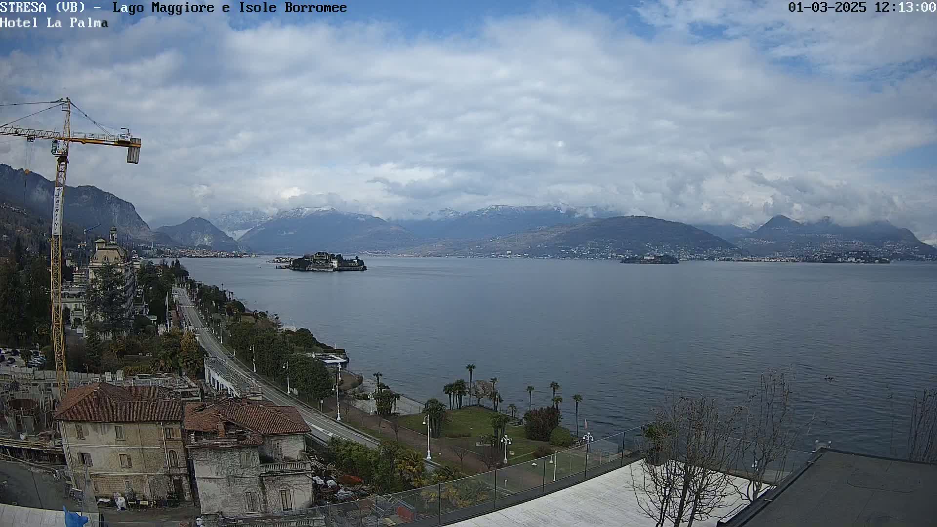 A partly cloudy day overlooks a calm lake with snow-capped mountains in the distance, a waterfront road, and buildings along the shore.