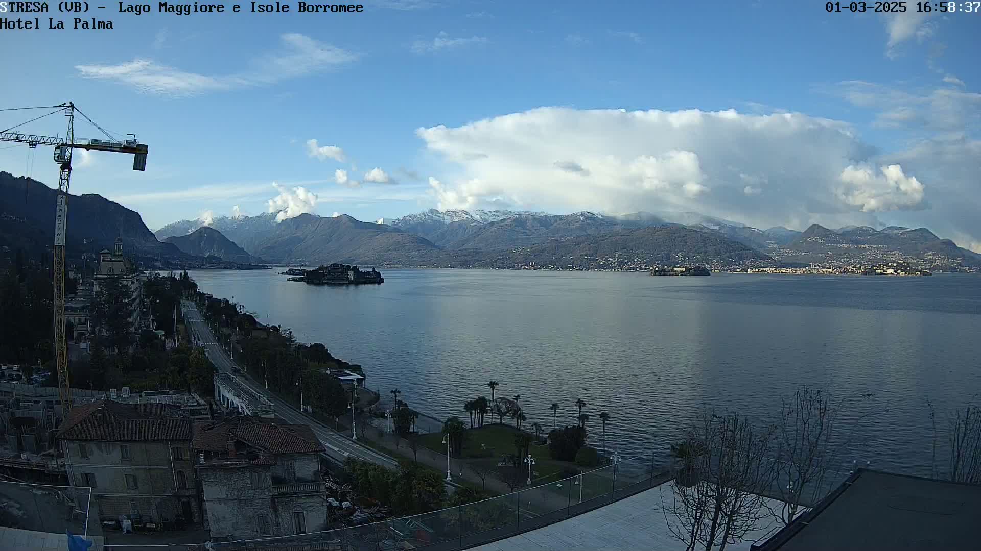 A partly cloudy day reveals a lake with snow-capped mountains in the background, a small island in the middle, and a construction crane near buildings along the shore.