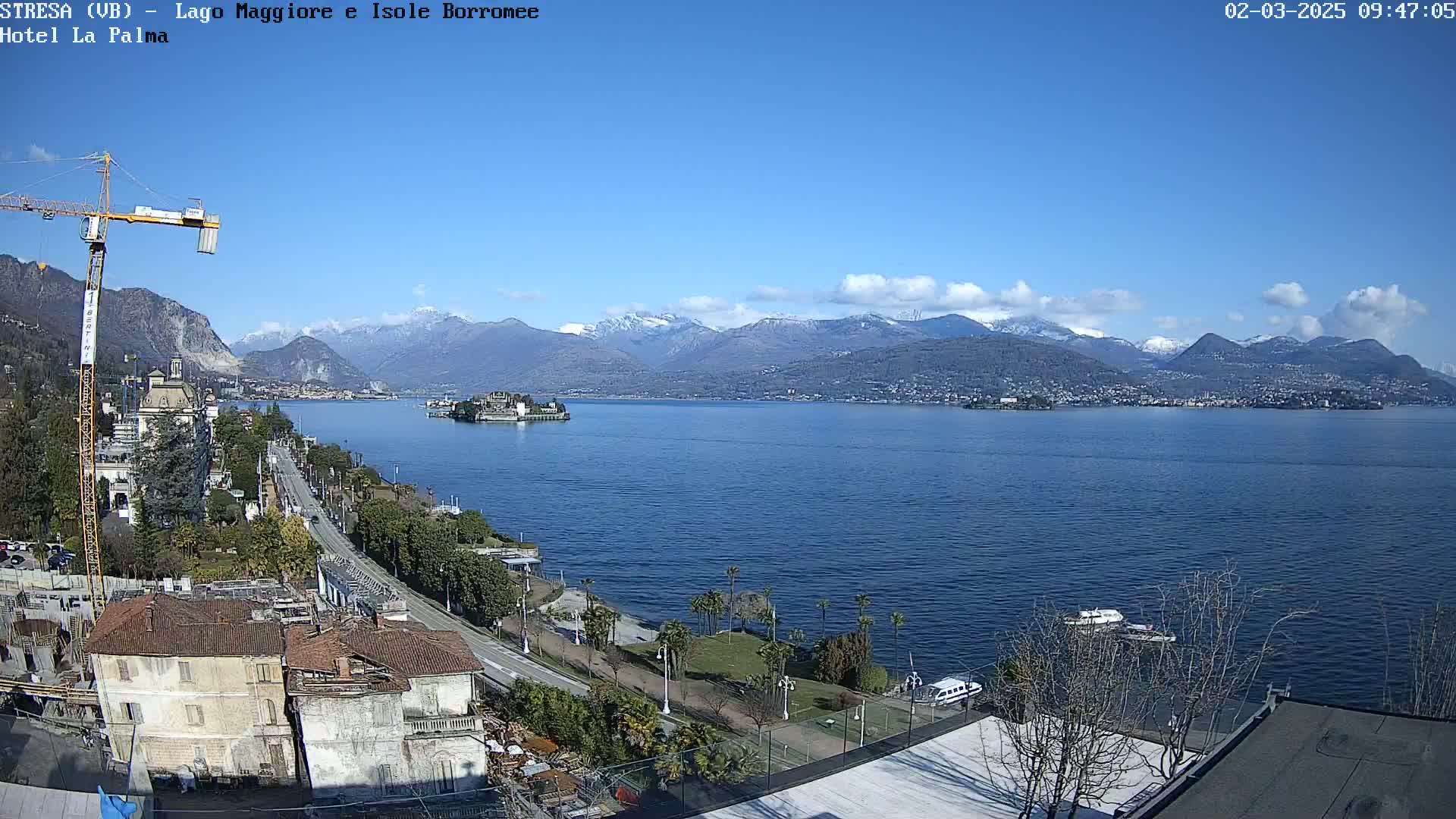 A clear, sunny day reveals a lake with snow-capped mountains in the background, a town on the lakeshore, and a construction crane near buildings under construction.