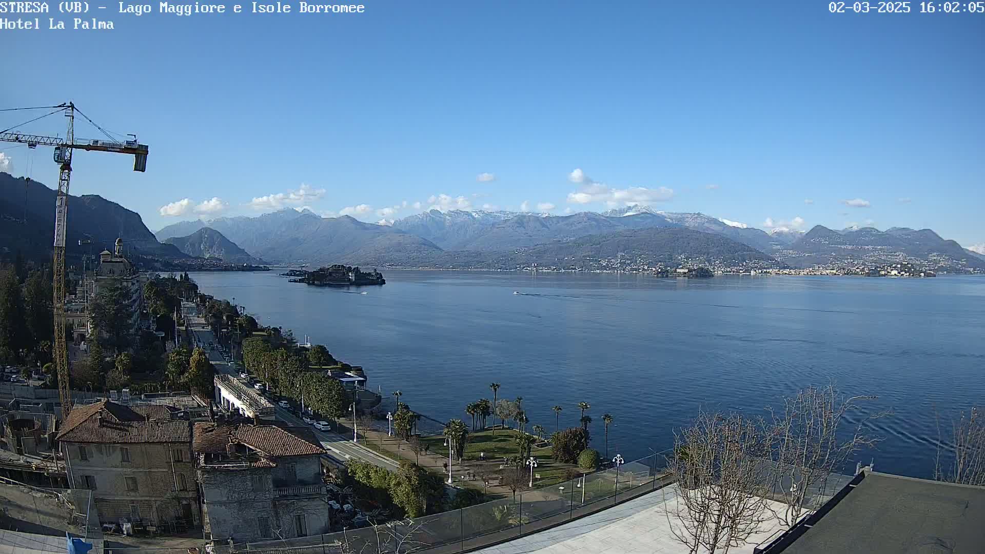 A clear, sunny day reveals a large lake with snow-capped mountains in the background, a small island in the middle of the lake, and a waterfront town with palm trees and a construction crane.