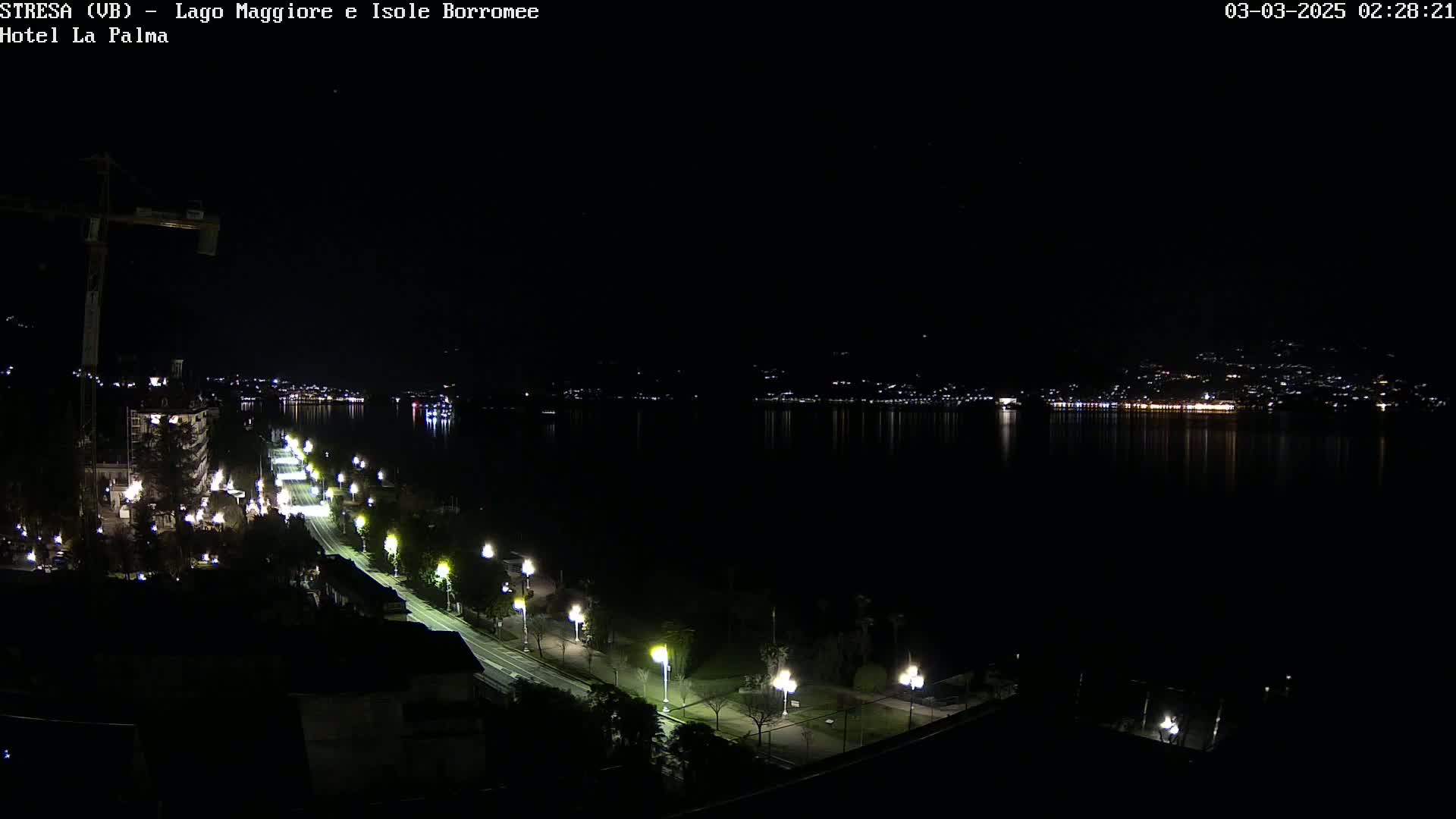 A nighttime view of a city's waterfront, featuring a road lined with lights leading to a dark body of water reflecting the distant city lights.