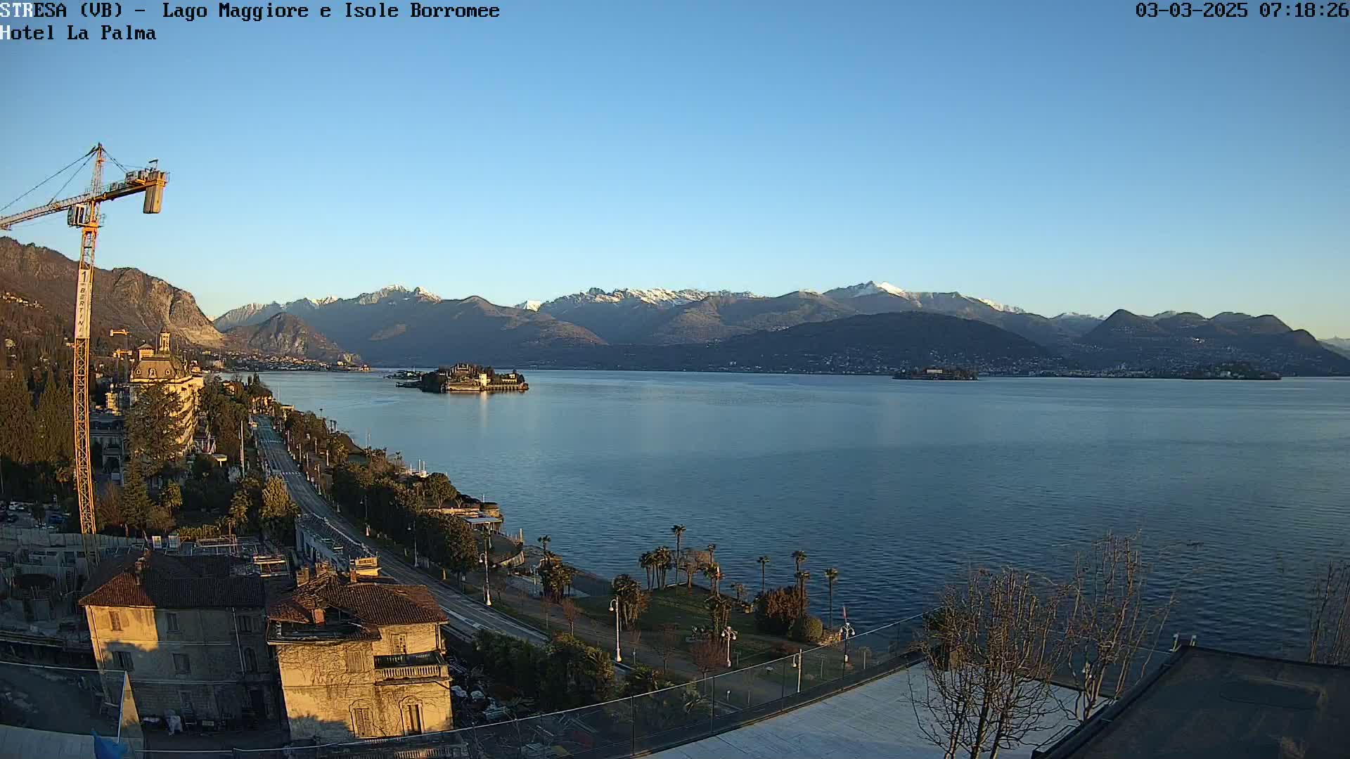 A clear, sunny day reveals a large lake with snow-capped mountains in the background, a small island with a building in the middle of the lake, and a coastal road lined with buildings.