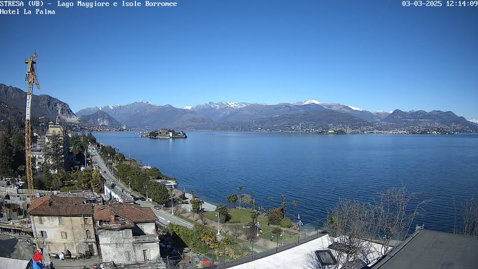 A sunny view of a lake with snow-capped mountains in the background, a town along the shoreline, and a small island in the lake.