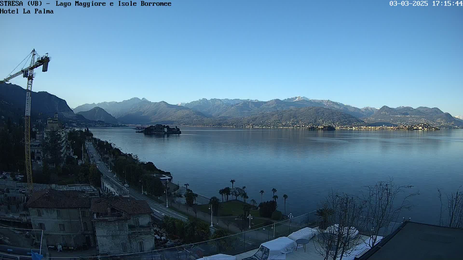 A clear, sunny day reveals a calm lake, bordered by a town, a small island, and snow-capped mountains in the background.