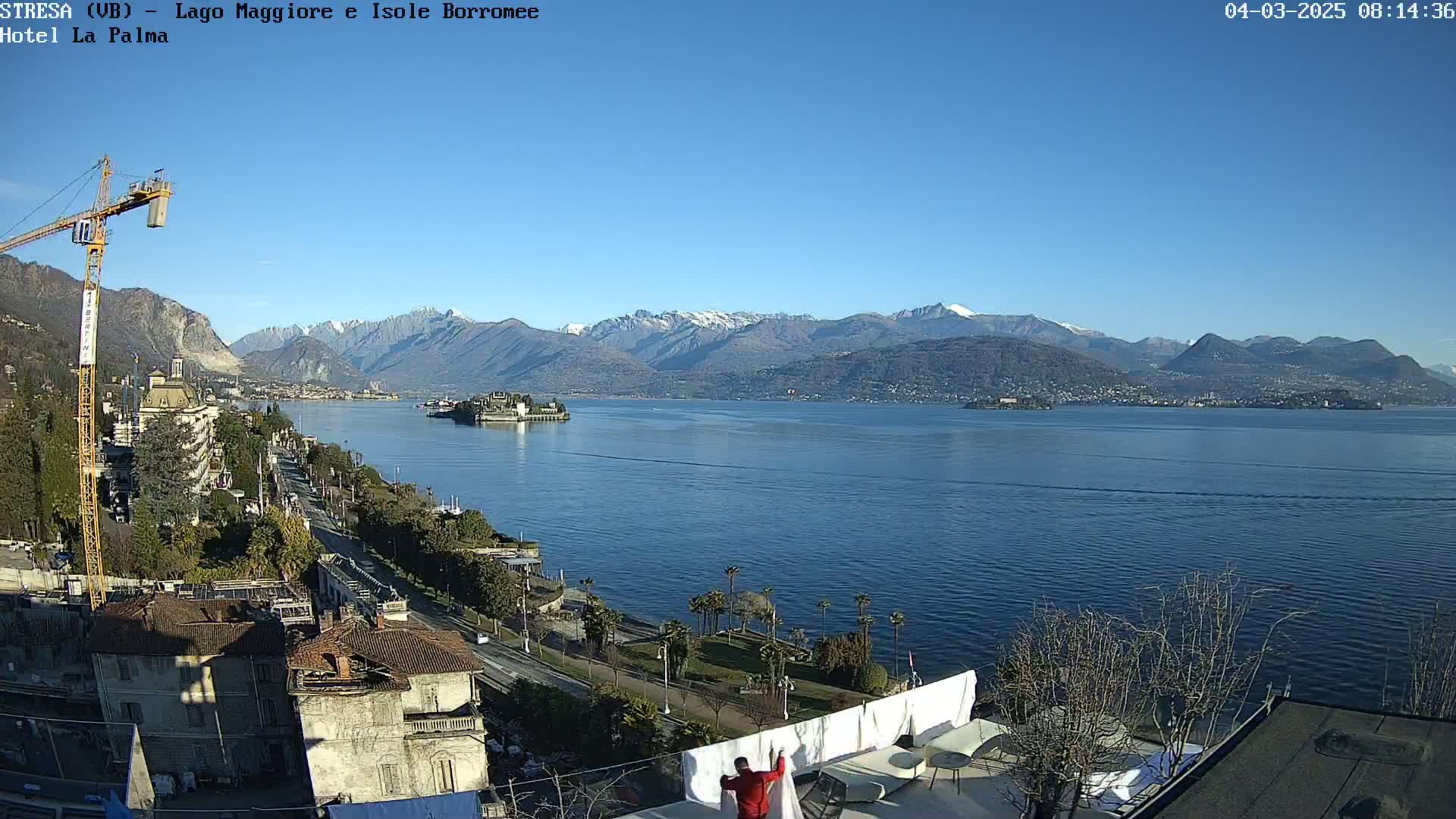 A clear, sunny day overlooks a large lake with snow-capped mountains in the background, a small island in the middle of the lake, and a cityscape with a crane visible along the shore.