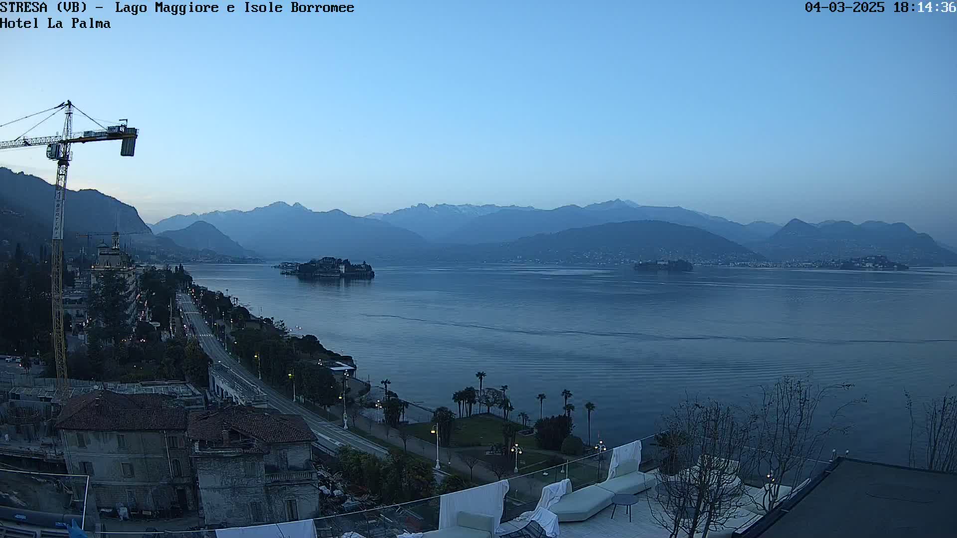 A twilight view overlooking a calm lake, mountains in the distance, a shoreline with buildings and palm trees, and a rooftop patio with lounge chairs.