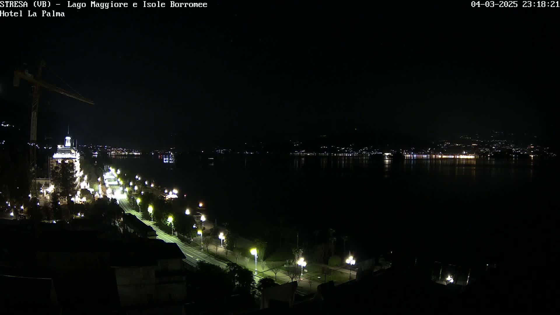 A nighttime view of a city street lined with lit lampposts, leading to a dark body of water reflecting the lights of buildings across the shore.
