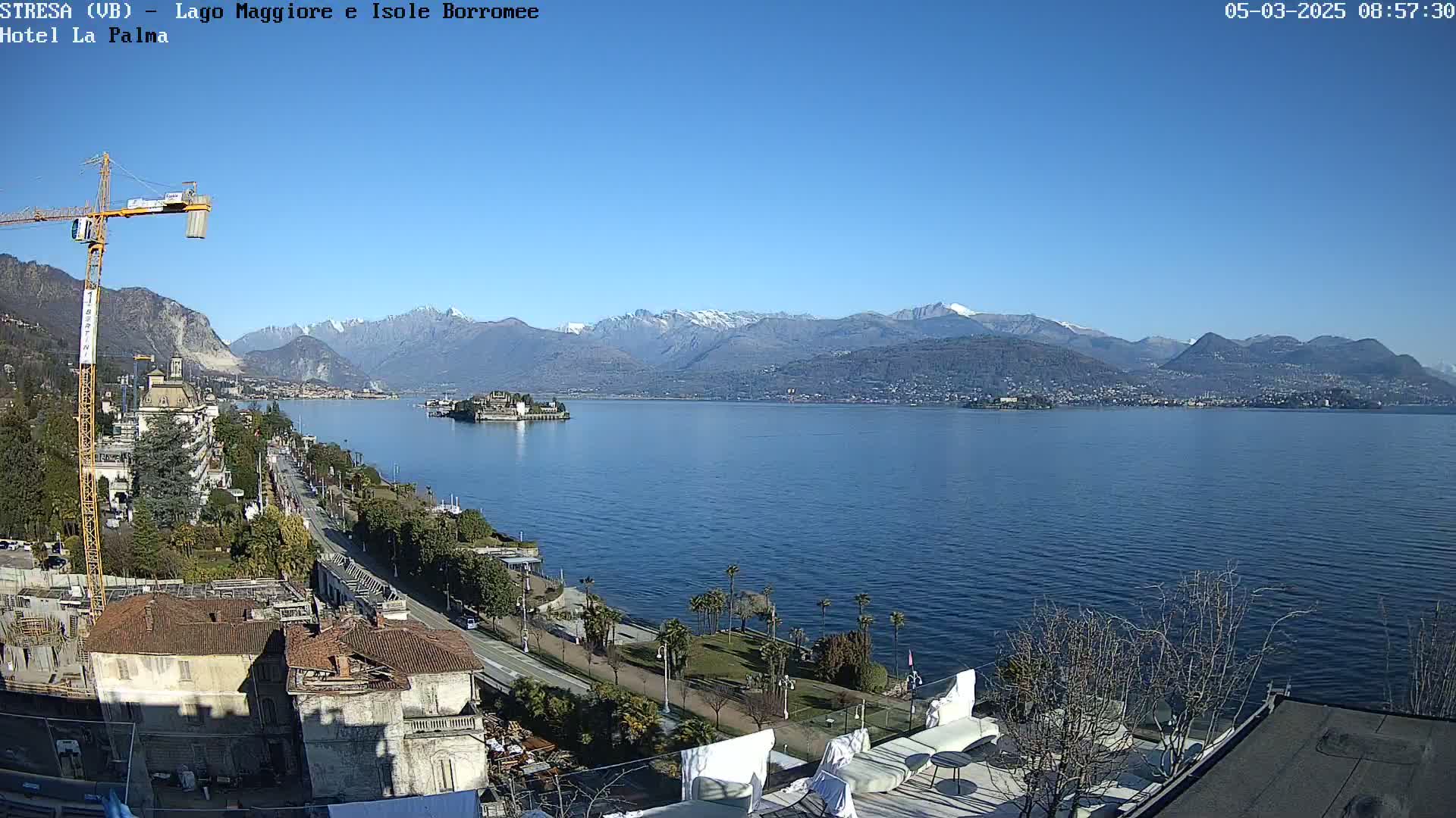 A sunny day overlooking a calm lake with snow-capped mountains in the background, a town along the lakeshore, and a construction crane near the foreground.