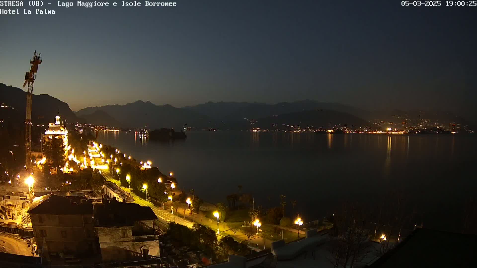 A nighttime view of a lakeside town, illuminated by streetlights, with a calm lake reflecting the lights of buildings across the water and mountains in the background under a dark sky.