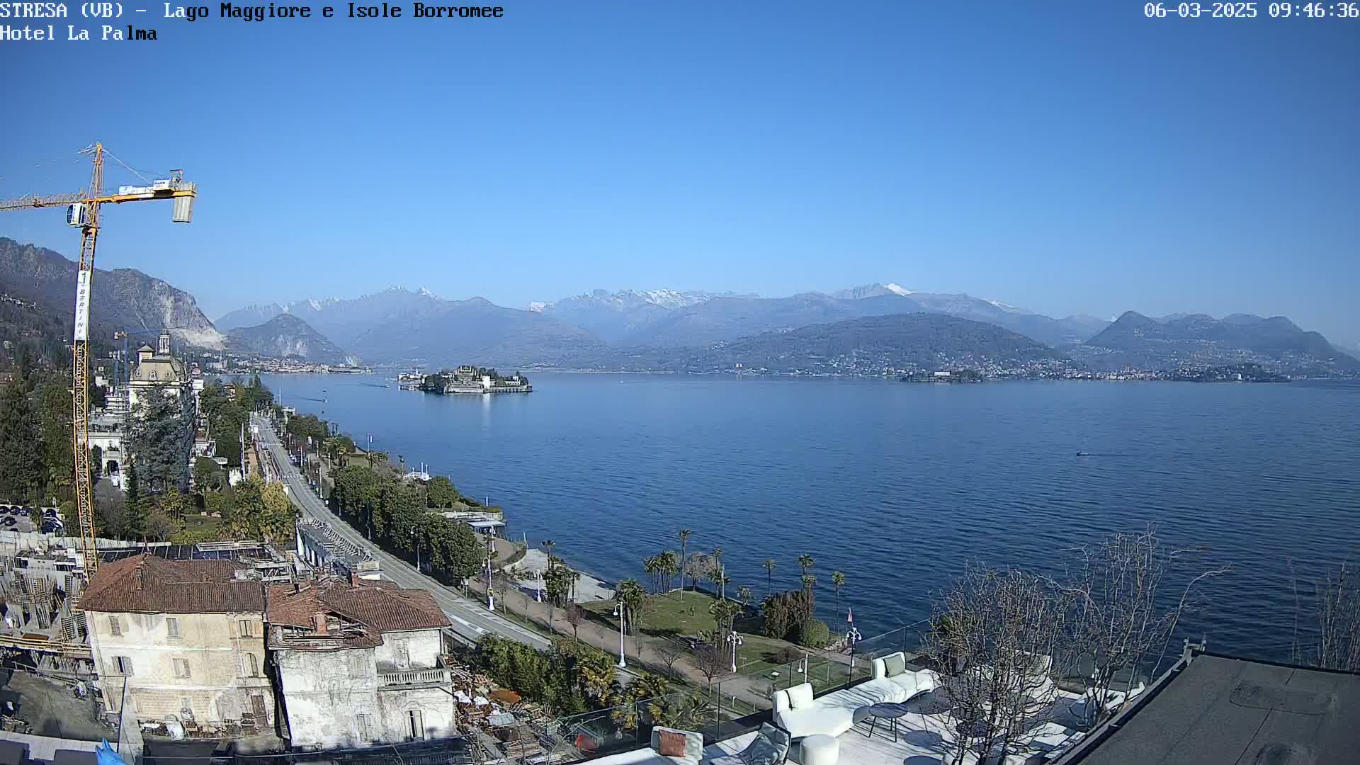 A clear, sunny day overlooking a large lake with snow-capped mountains in the background, a town and island visible on the lake, and a construction crane visible in the foreground.