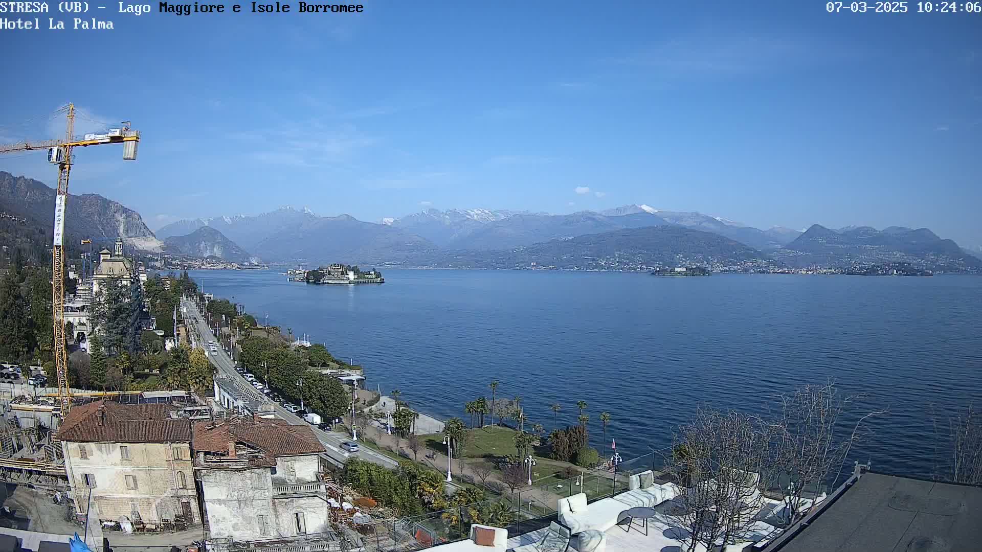 A sunny day overlooking a lake with snow-capped mountains in the distance, a town along the shoreline, and a construction crane in the foreground.