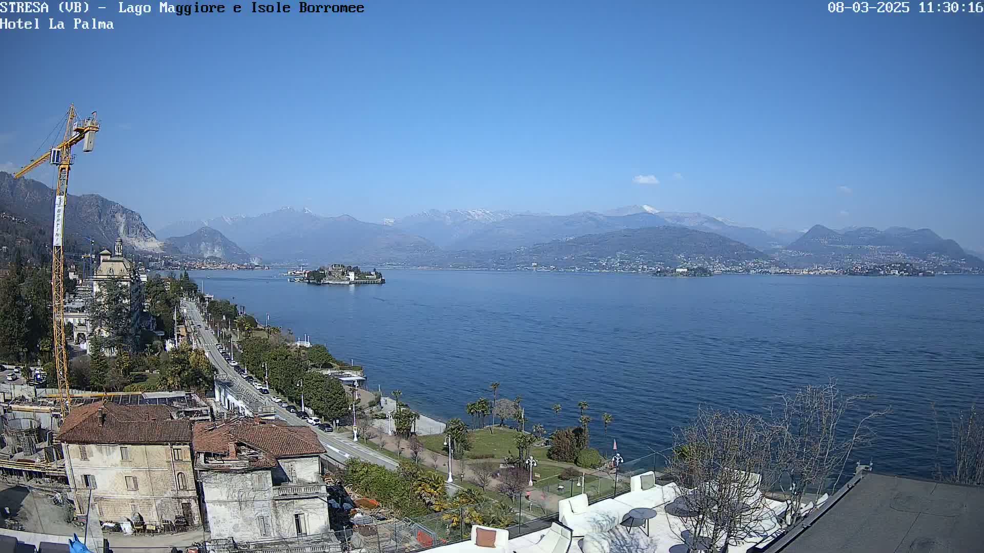 A sunny day reveals a large body of water, with a small island in the middle distance, buildings and a construction crane close by, and snow-capped mountains in the background.
