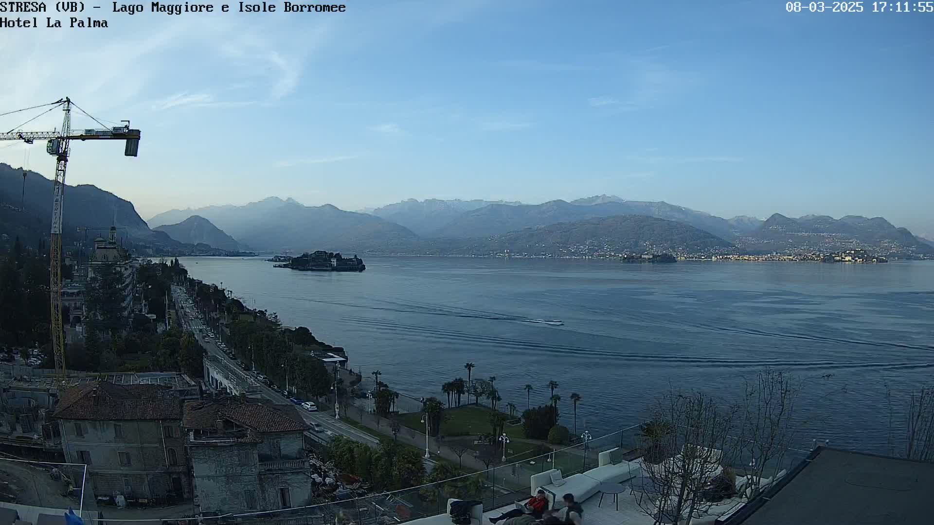 A clear, daytime view of a lake, with mountains in the background, a small island in the middle of the lake, a construction crane to the left, and buildings and palm trees along the lakeshore.