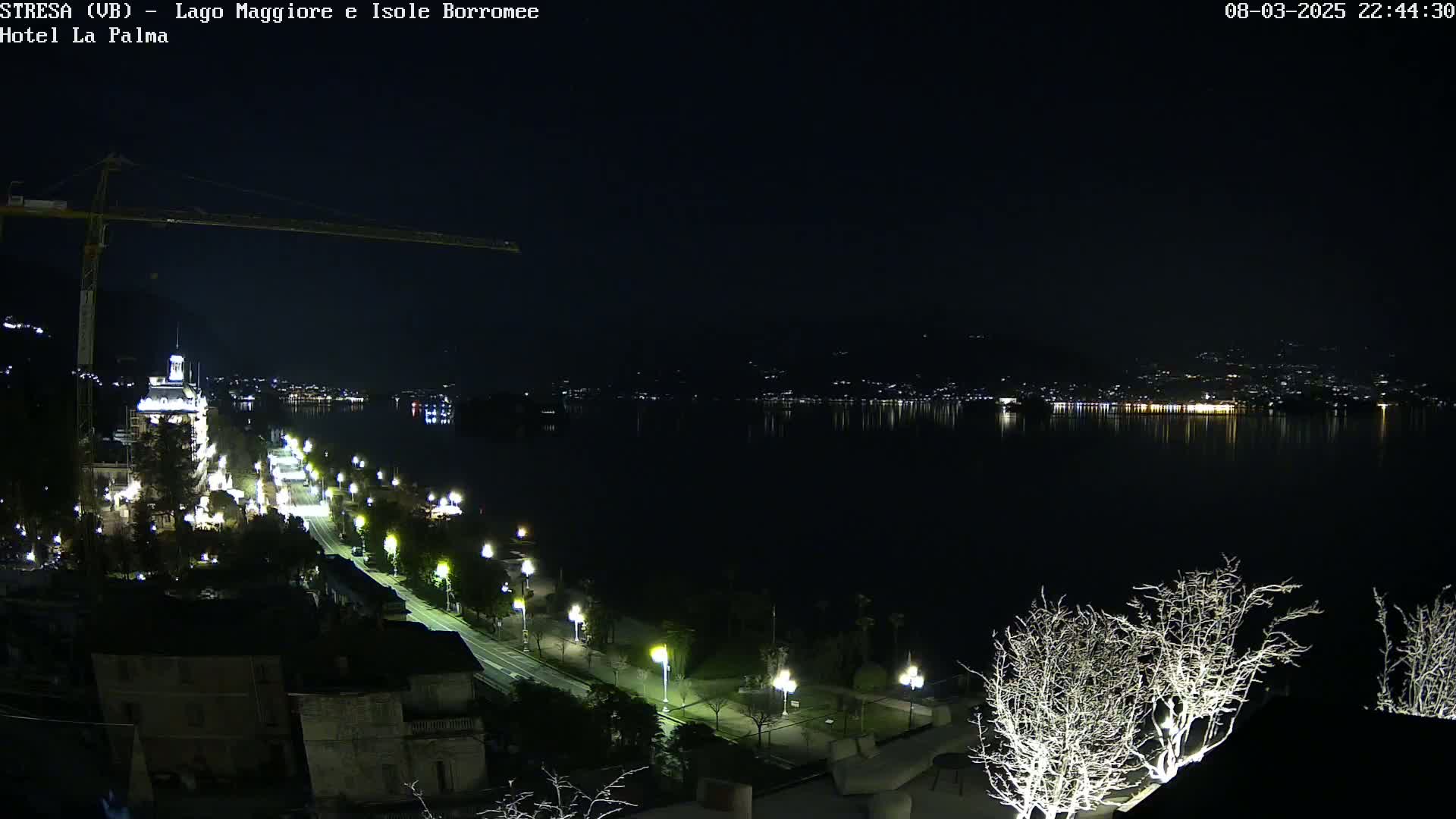 A nighttime view of a city's waterfront, featuring a calm lake reflecting lights from buildings across the water, a road lined with lights, and a construction crane in the foreground.