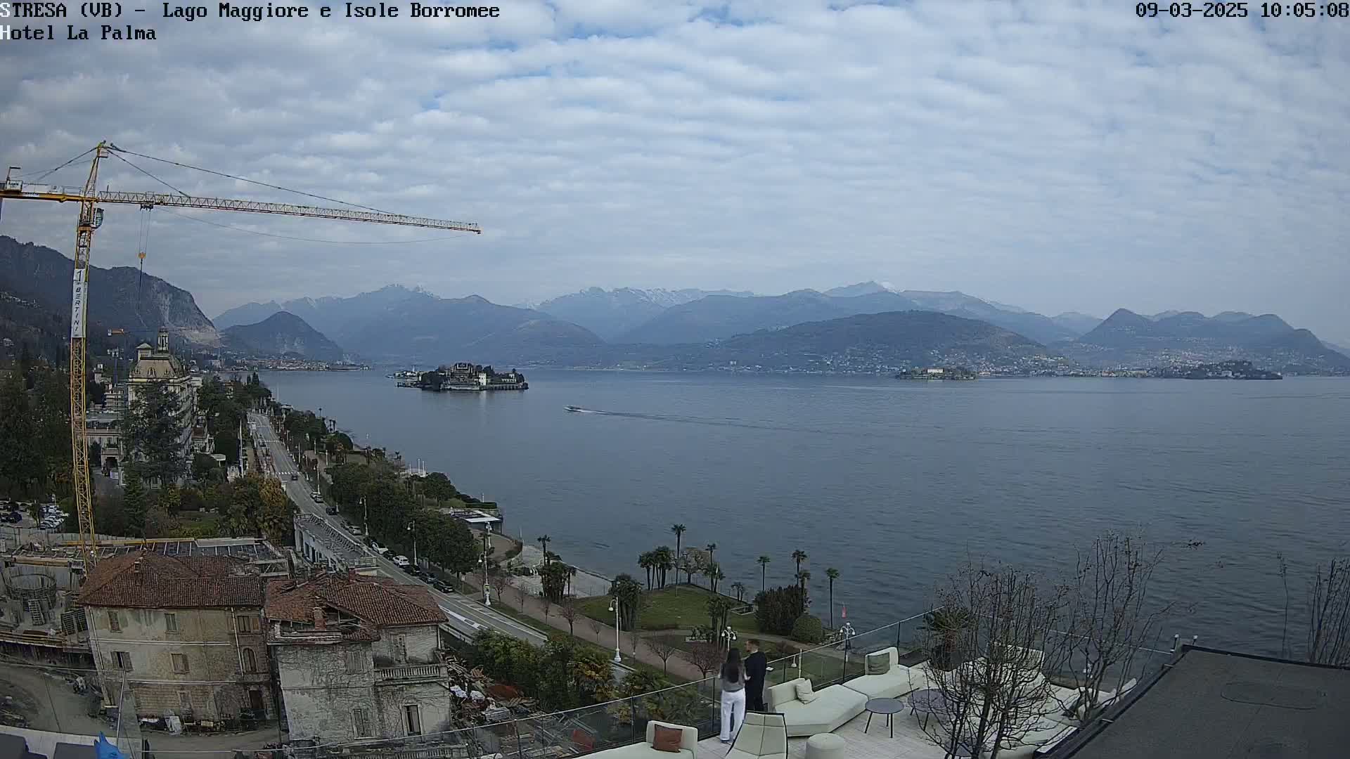 A partly cloudy day overlooks a large lake with mountains in the background, a crane, buildings, and a small boat are visible near the shore.