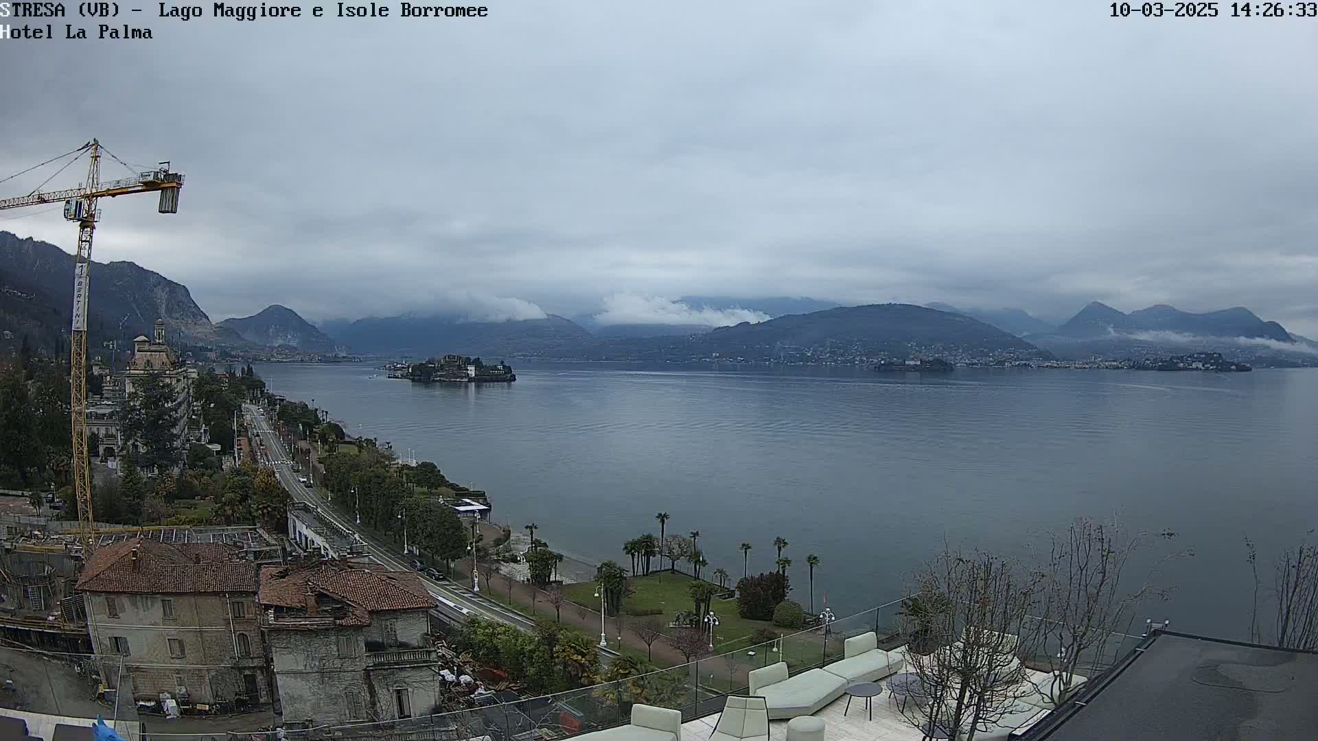 A large body of water is seen from a high vantage point, with mountains in the background partially obscured by fog under an overcast sky; a crane, buildings, and trees are visible in the foreground.