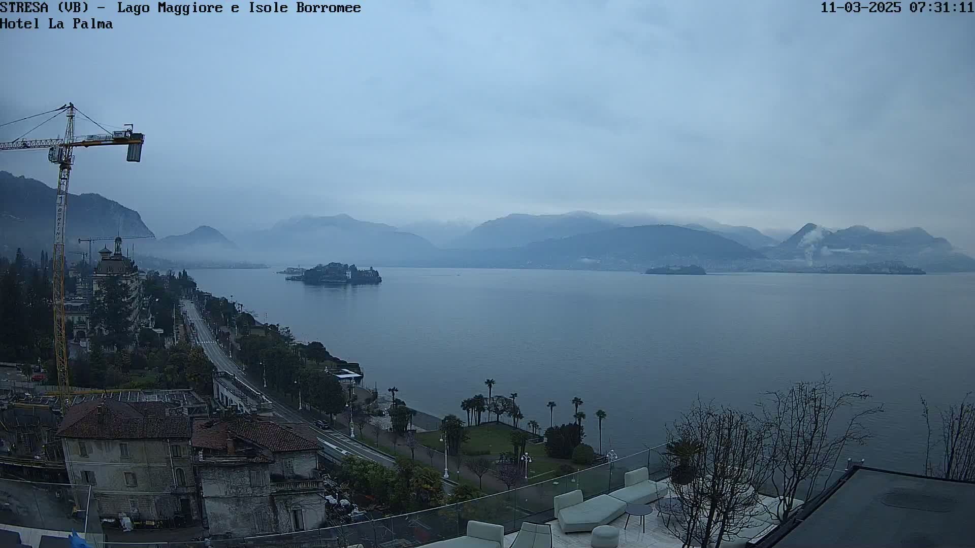 A hazy, overcast day reveals a large lake surrounded by mountains, with a town and a construction crane visible in the foreground.