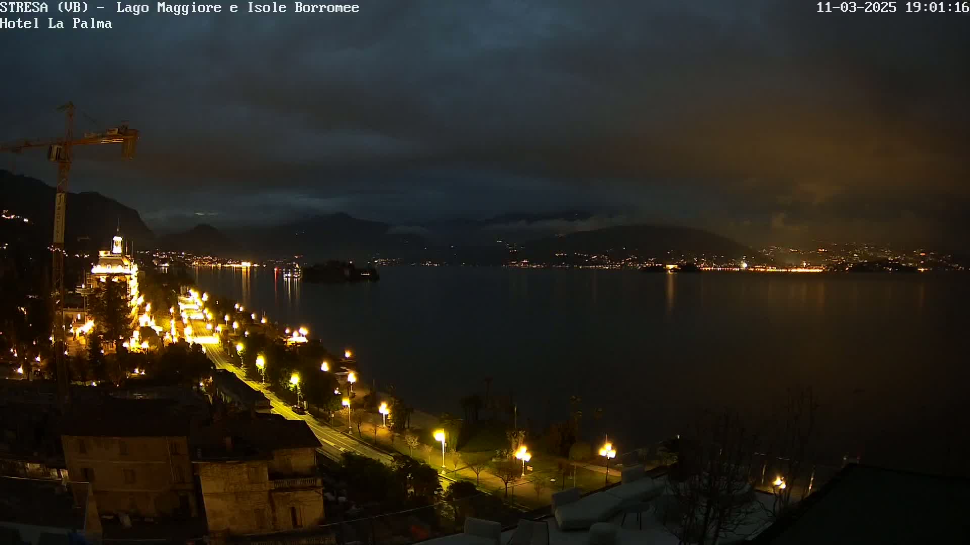A nighttime view of a city along a lake, illuminated by streetlights, with a construction crane visible and a partly cloudy sky.