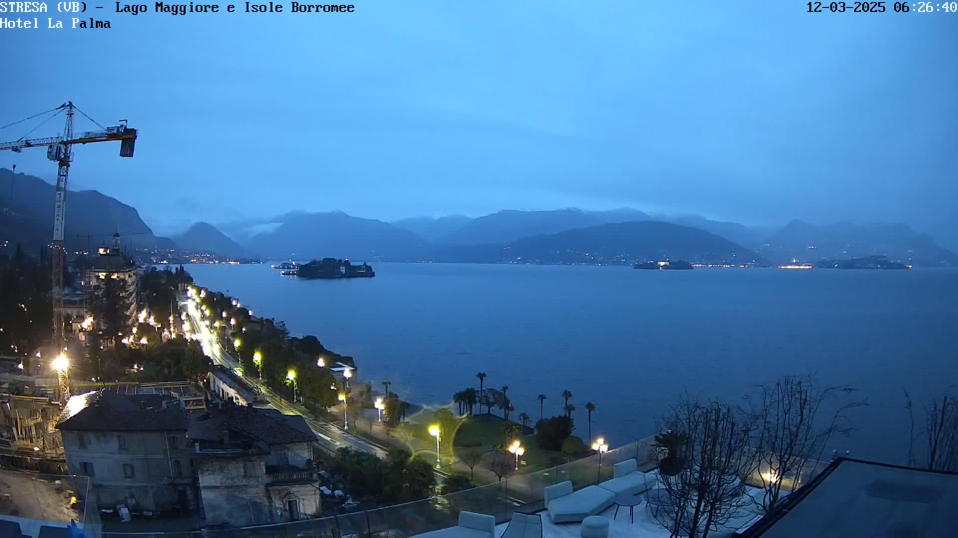 A nighttime, rain-soaked lakeside town with a large lake and mountains in the distance, illuminated by streetlights and the lights of buildings.