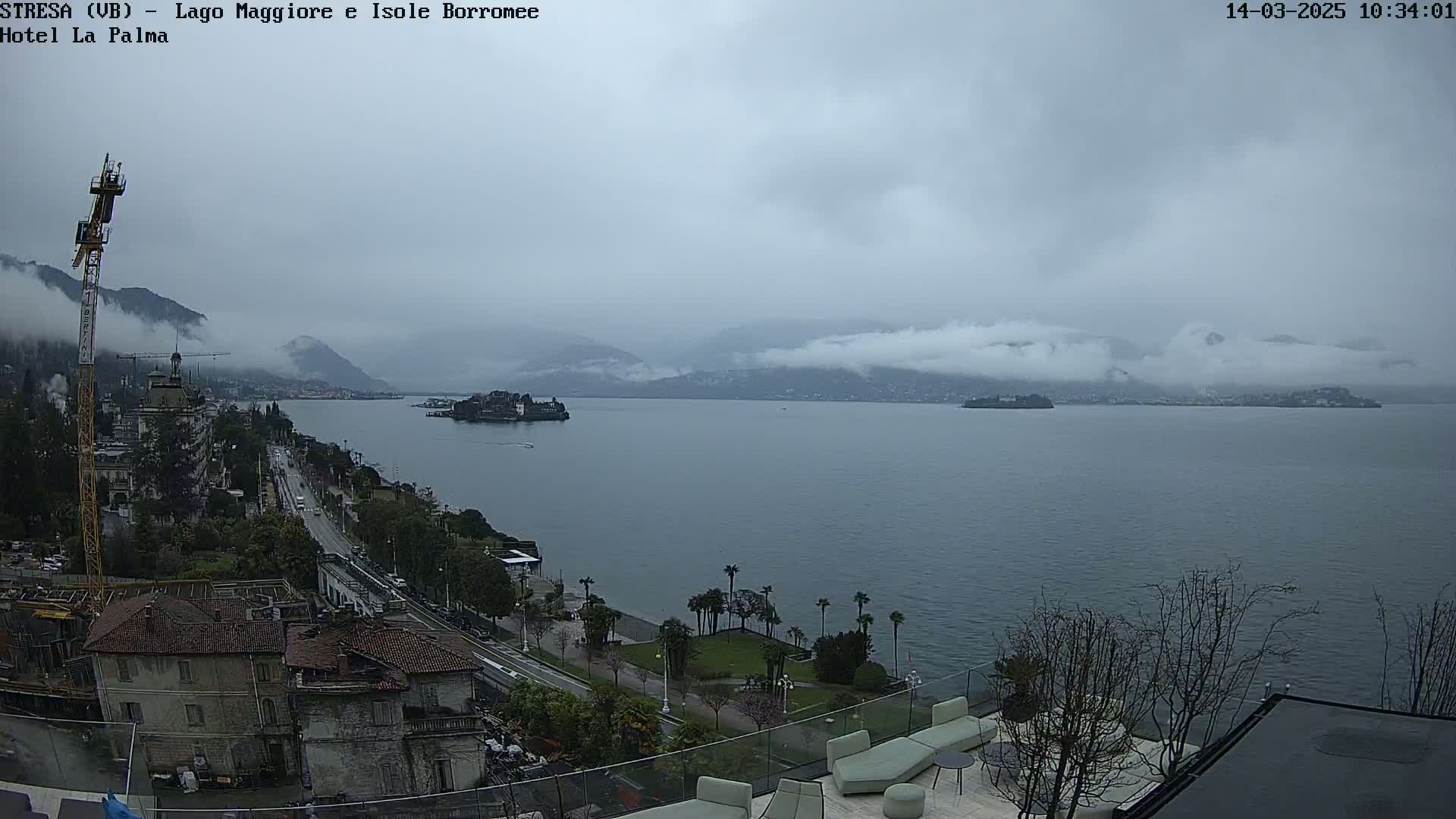 A cloudy, overcast day reveals a lake with islands and mountains in the distance, seen from an elevated vantage point overlooking a waterfront road and buildings.