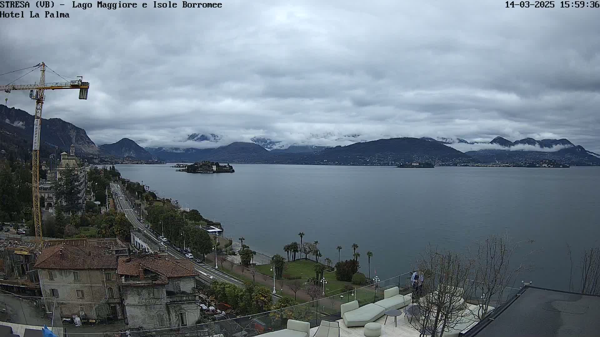 A large lake is seen from an elevated vantage point on an overcast day, with mountains in the background partially obscured by clouds, and a shoreline with buildings and vegetation in the foreground.