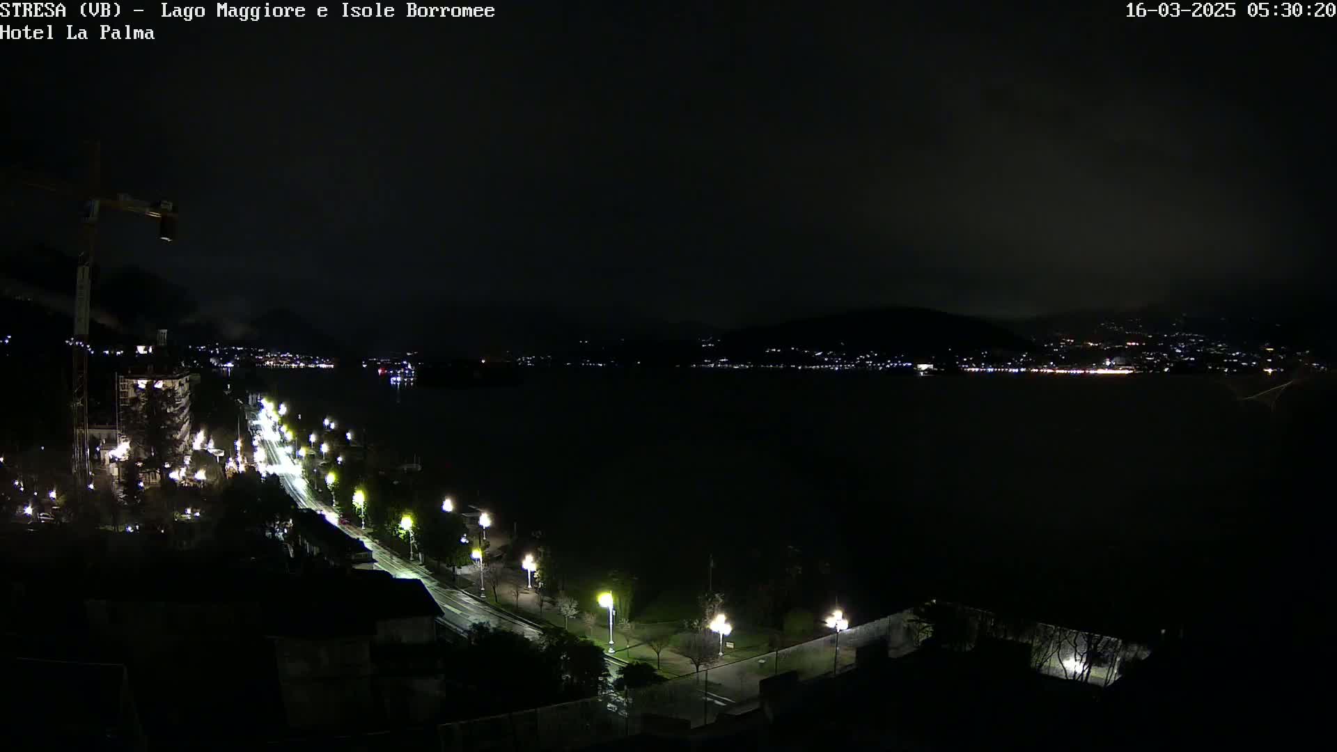 A nighttime view of a dark lake, a city's lights on the opposite shore, and a road lined with streetlights leading toward the water.