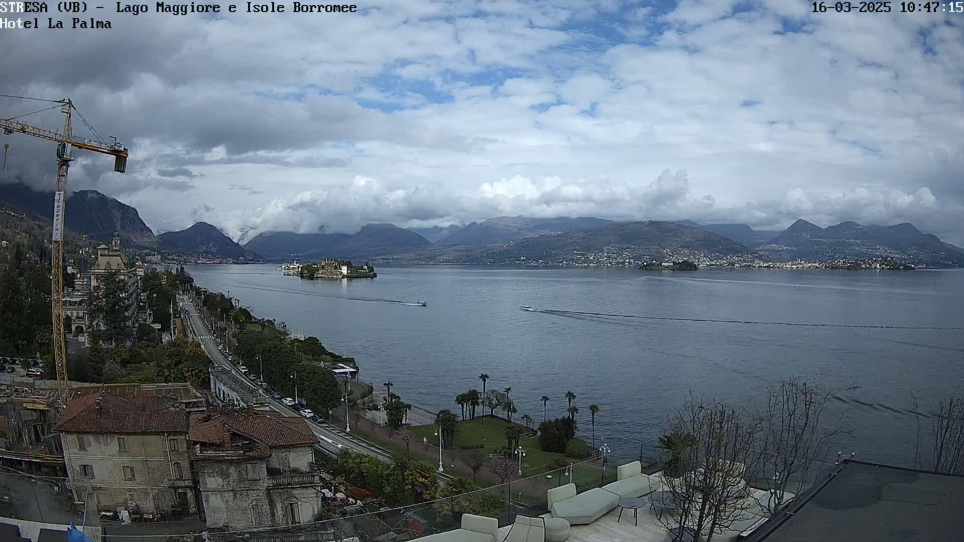 A partly cloudy day overlooking a lake with mountains in the distance, a town along the shore, and a construction crane in the foreground.