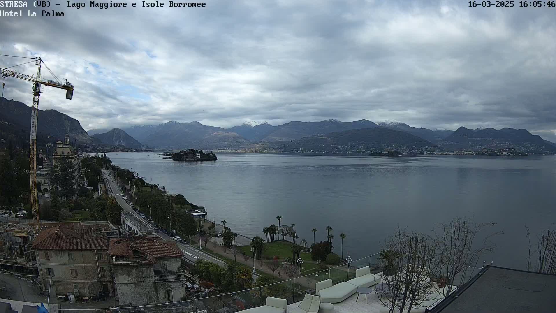 A view of a lake surrounded by mountains with some snow, under a cloudy sky, from a high vantage point showing a town, a construction crane, and a terrace with outdoor furniture.
