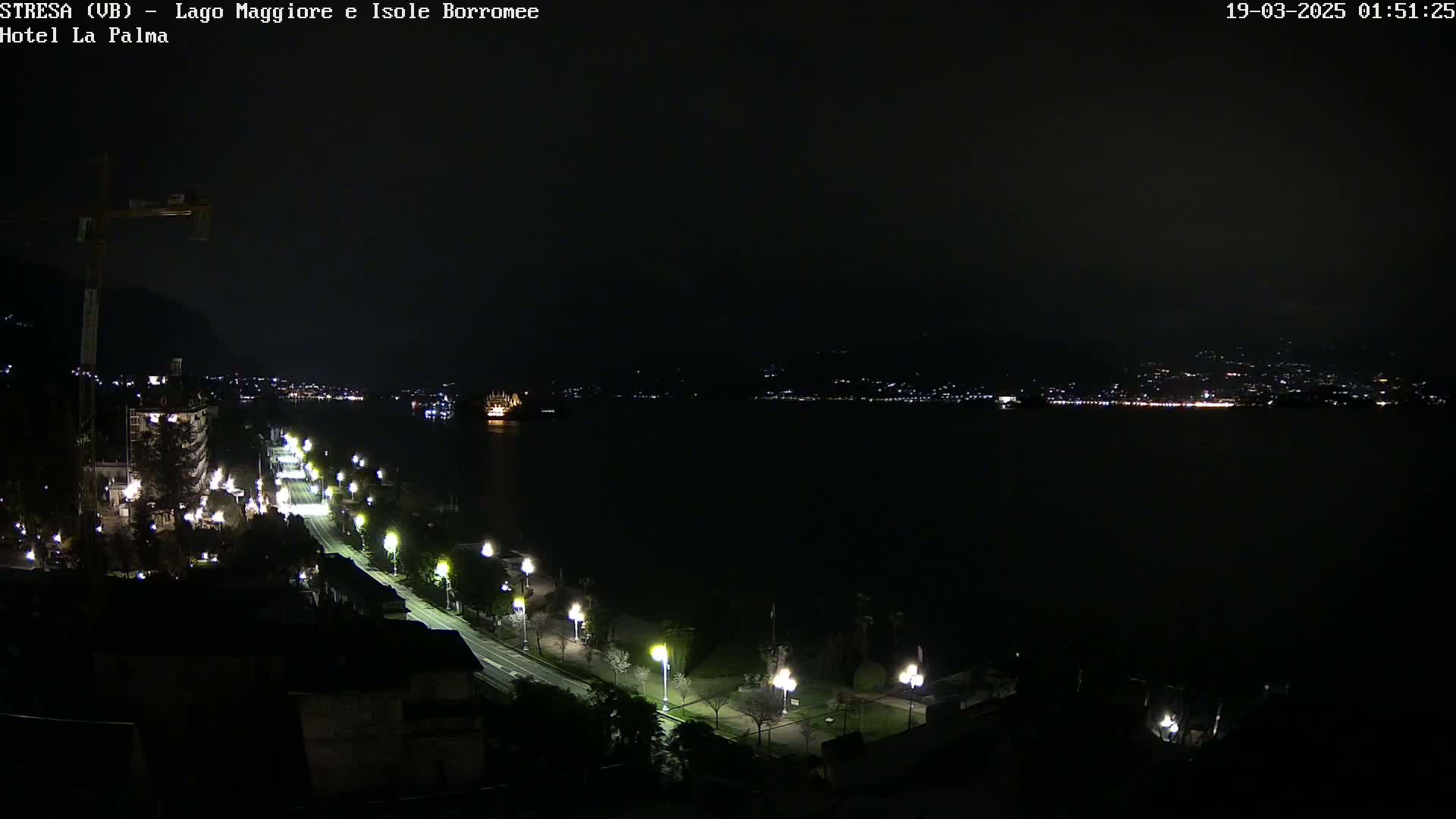 A nighttime, high-angle view shows a dark body of water with a line of lights on the opposite shore, a road with streetlights leading toward the water, and a construction crane visible in the upper left corner.