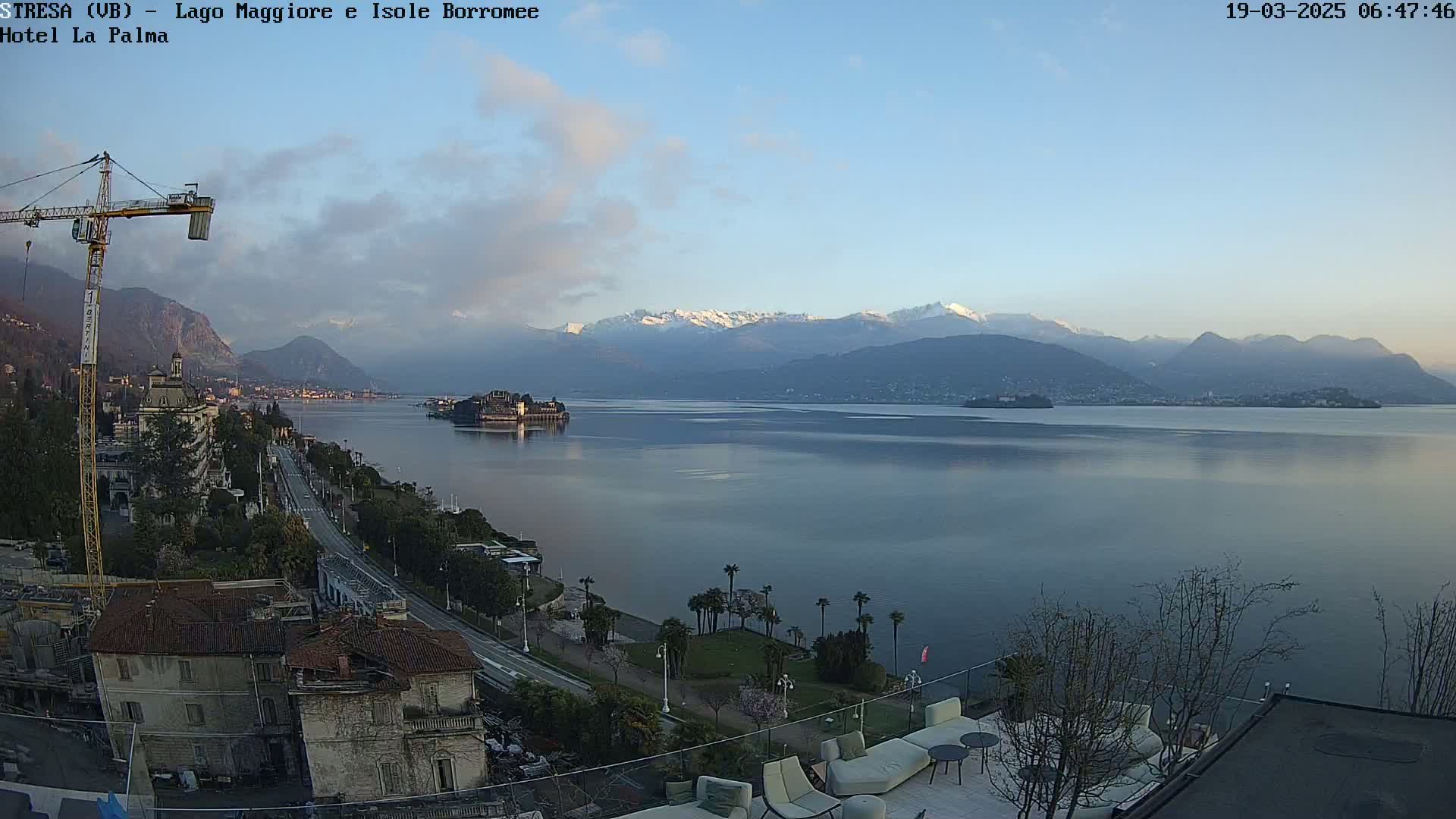 A calm lake with snow-capped mountains in the background is seen from a high vantage point, with a town and construction crane visible in the foreground under a clear, bright sky.
