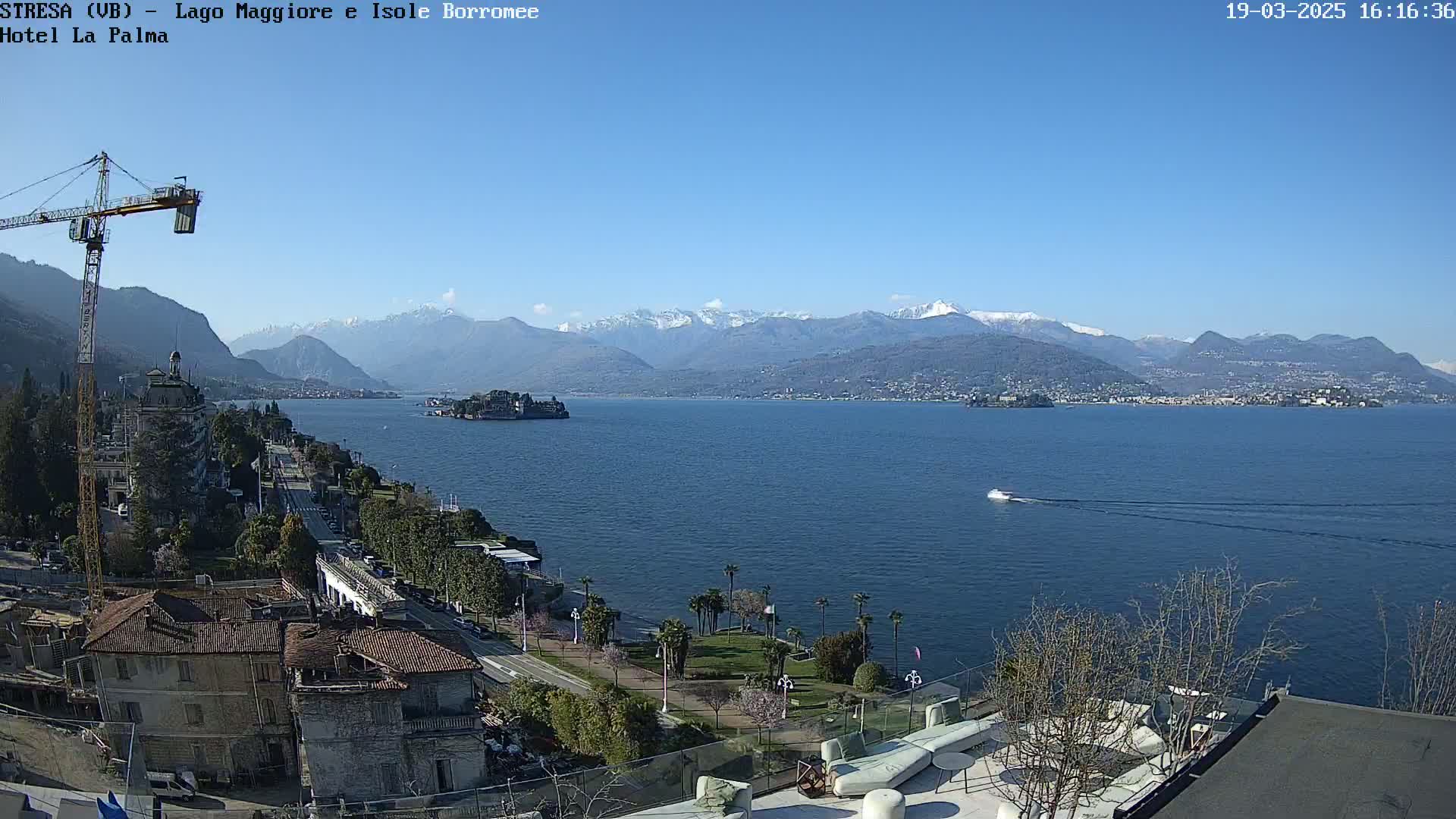 A clear, sunny day reveals a lake with snow-capped mountains in the background, a small island, and a town with buildings and a construction crane.