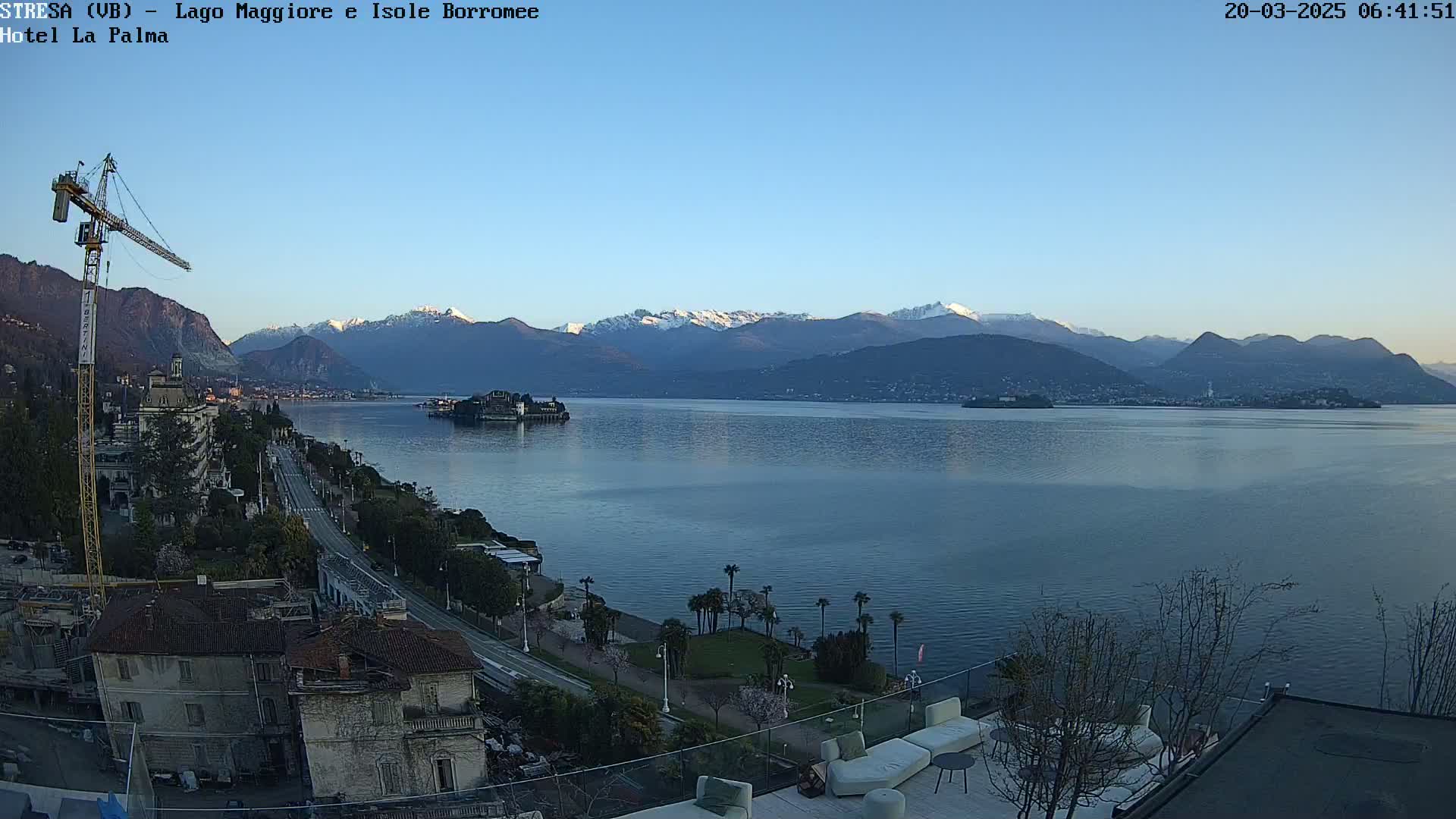 A calm lake stretches toward snow-capped mountains under a clear, light blue sky, with a town and a construction crane visible in the foreground.