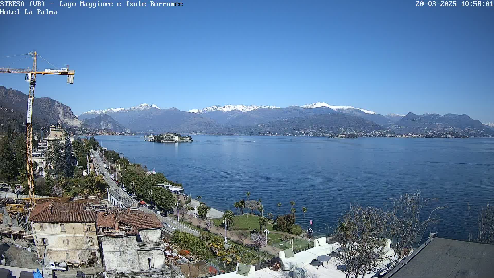 A clear, sunny day overlooks a lake with snow-capped mountains in the background, a shoreline with buildings and palm trees, and a construction crane visible near the foreground.