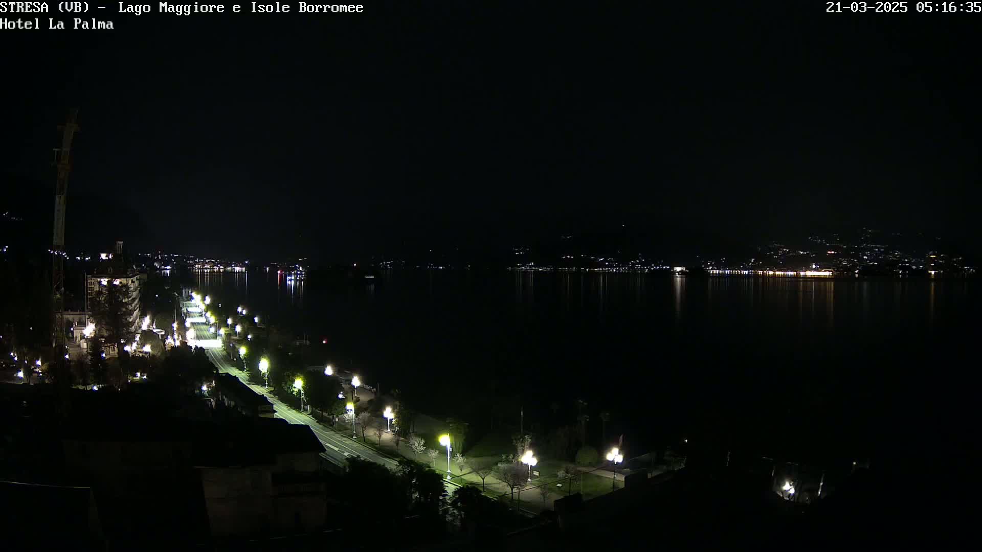 A nighttime view of a city's waterfront, featuring a dark lake reflecting city lights on the opposite shore, a road lined with illuminated lampposts, and buildings along the water's edge.