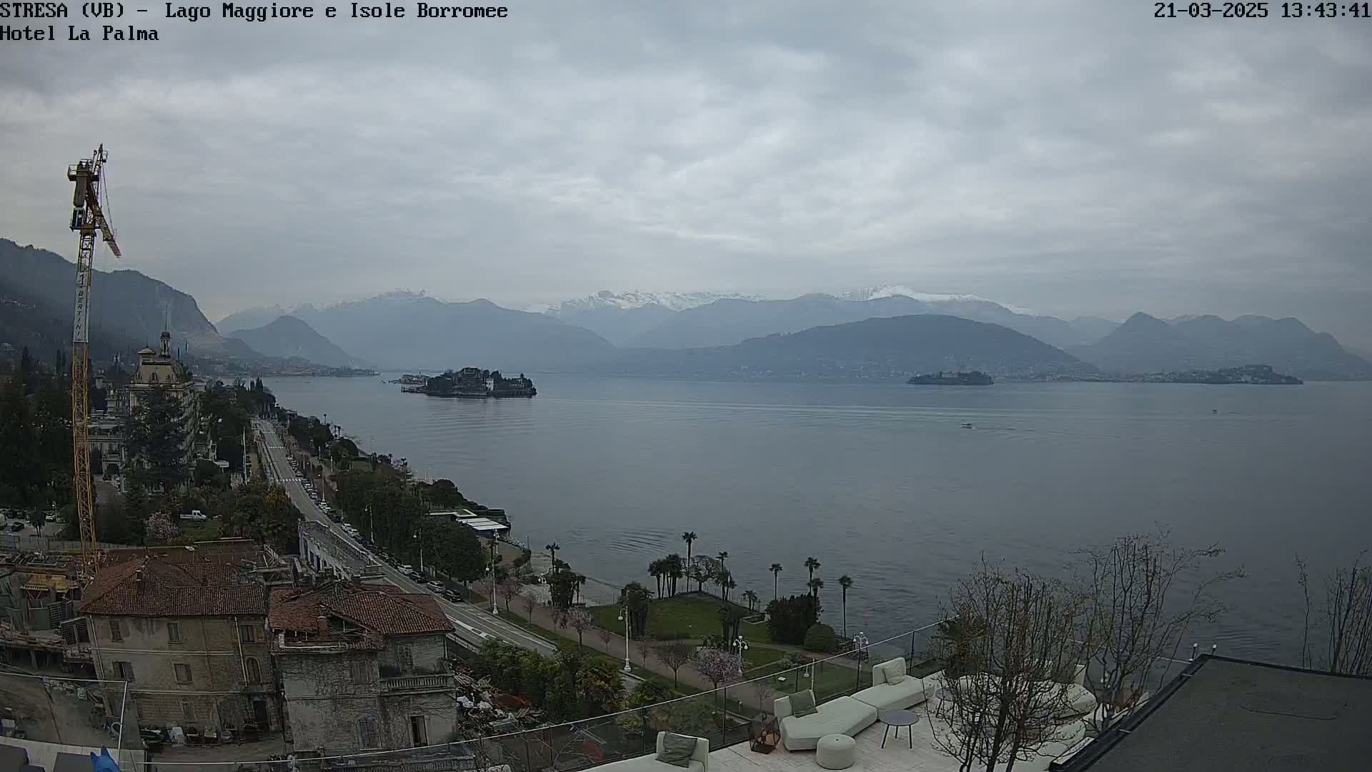 A large lake stretches out before a cityscape under a cloudy sky, with snow-capped mountains in the distance.