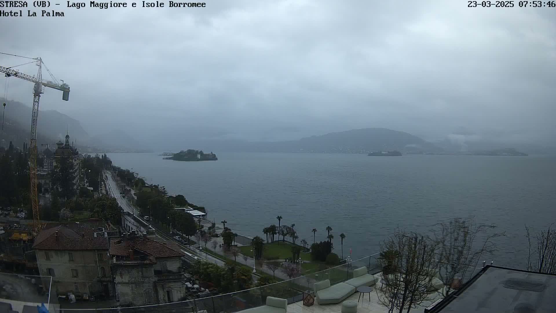 A rain-swept view of a lake, with a town and construction crane visible in the foreground, and distant hills under a cloudy sky.