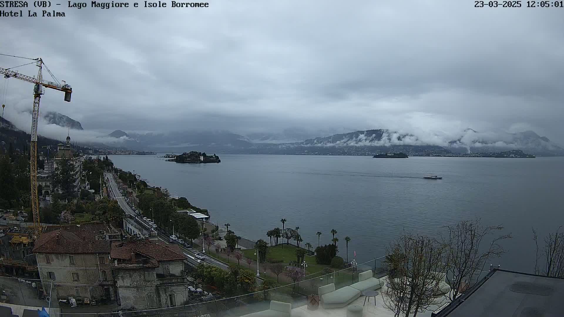 A cloudy day overlooks a large body of water with a small island and distant mountains partially shrouded in fog, from a high vantage point showing buildings and a road along the shore.