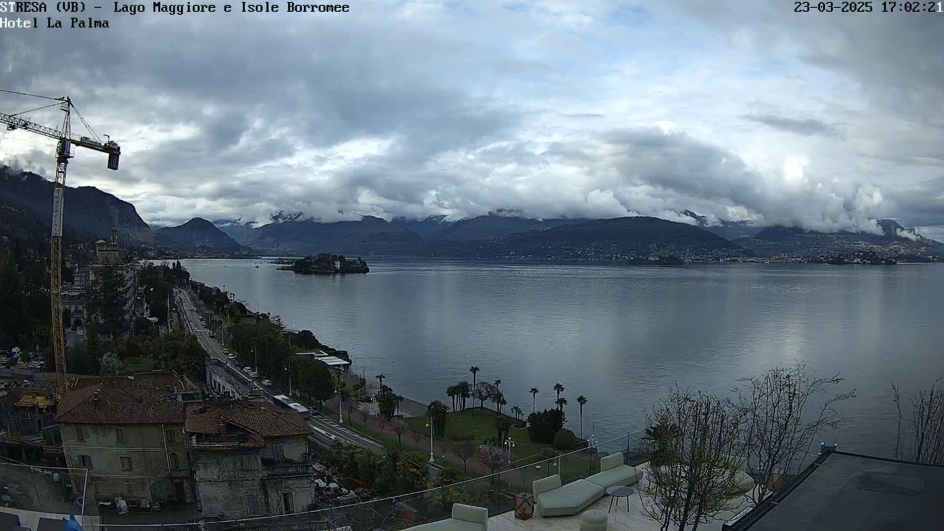 A cloudy day overlooks a large lake with mountains in the background, a small island visible in the middle distance, and a town with buildings and a roadway along the lakeshore.