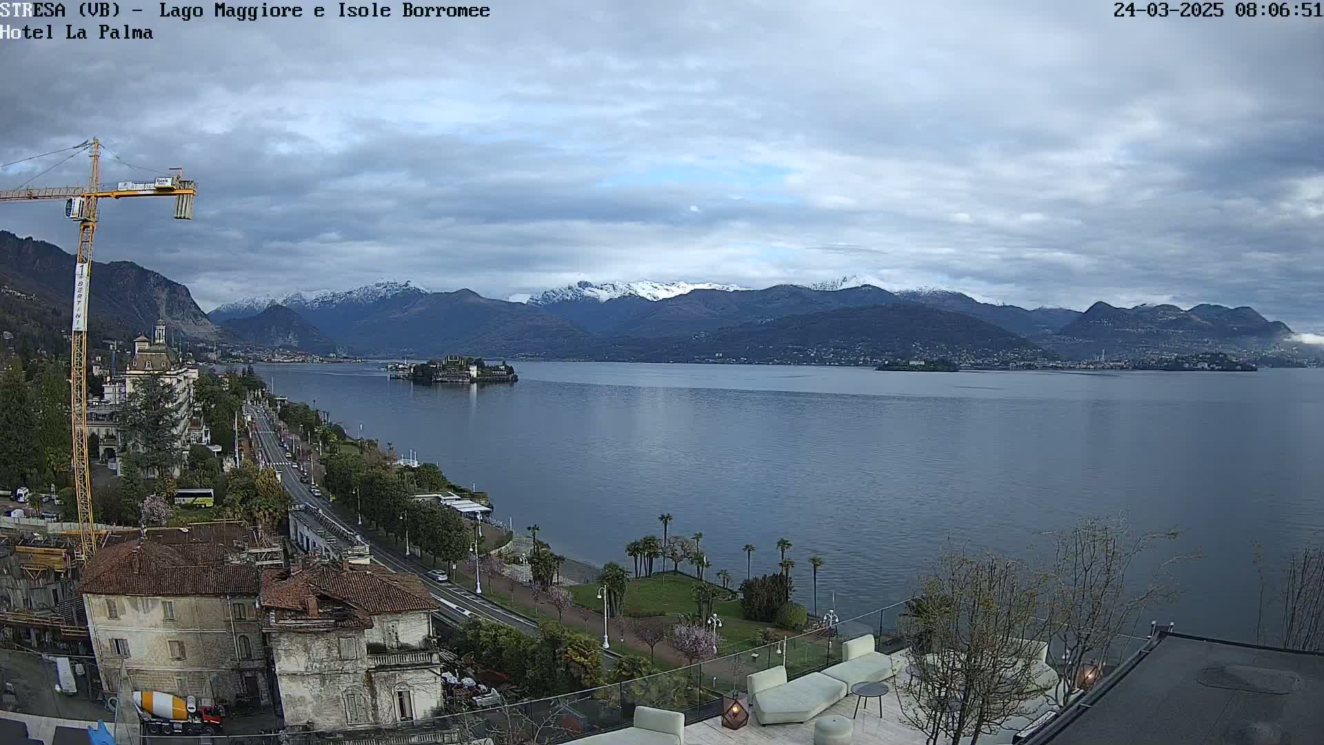A partly cloudy day reveals a lake with snow-capped mountains in the background, a town situated along the shoreline, and a construction crane in the foreground.