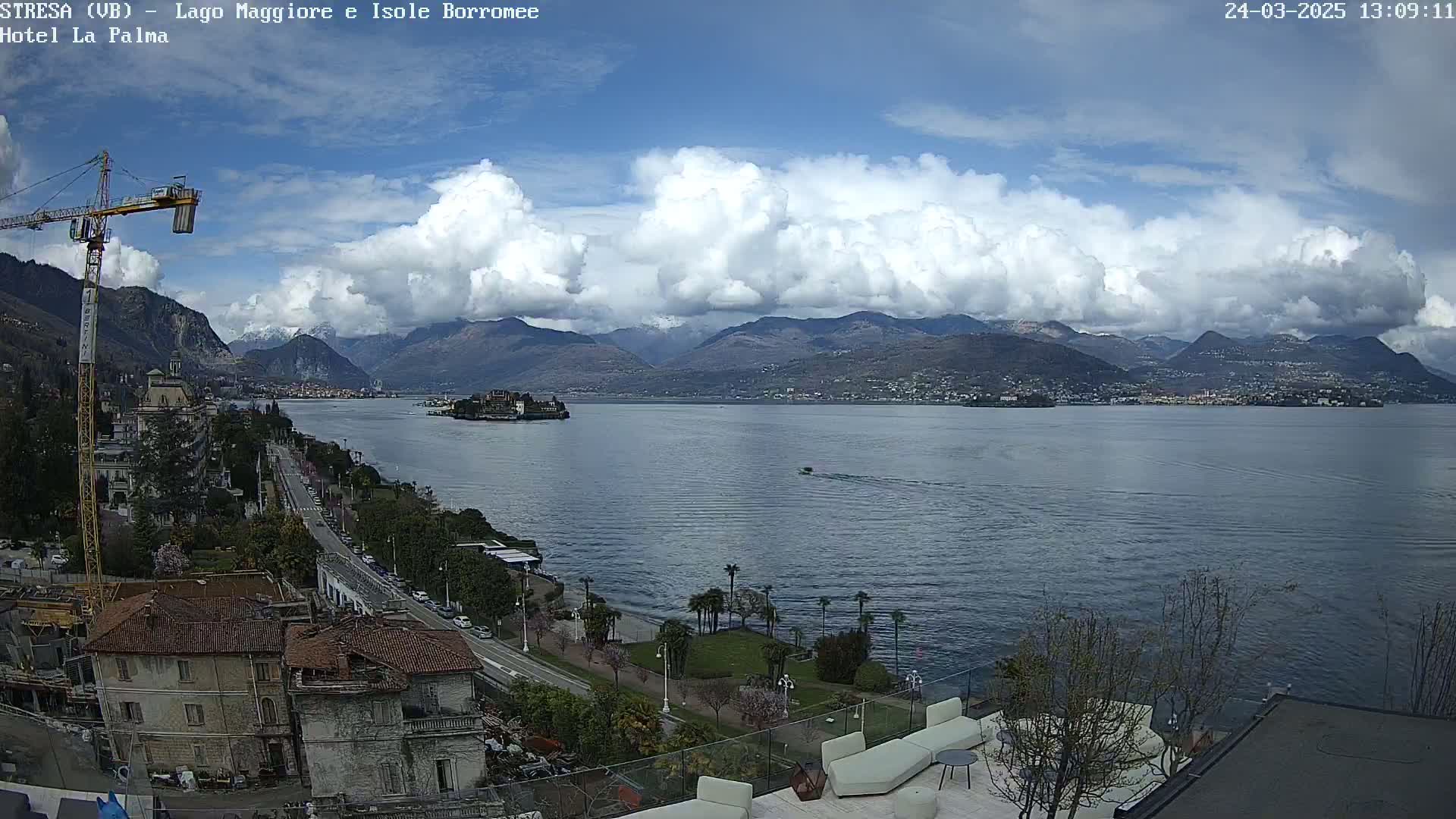 A partly cloudy day overlooks a large lake with mountains in the background, a small island in the middle of the lake, and a roadway and buildings along the shoreline.