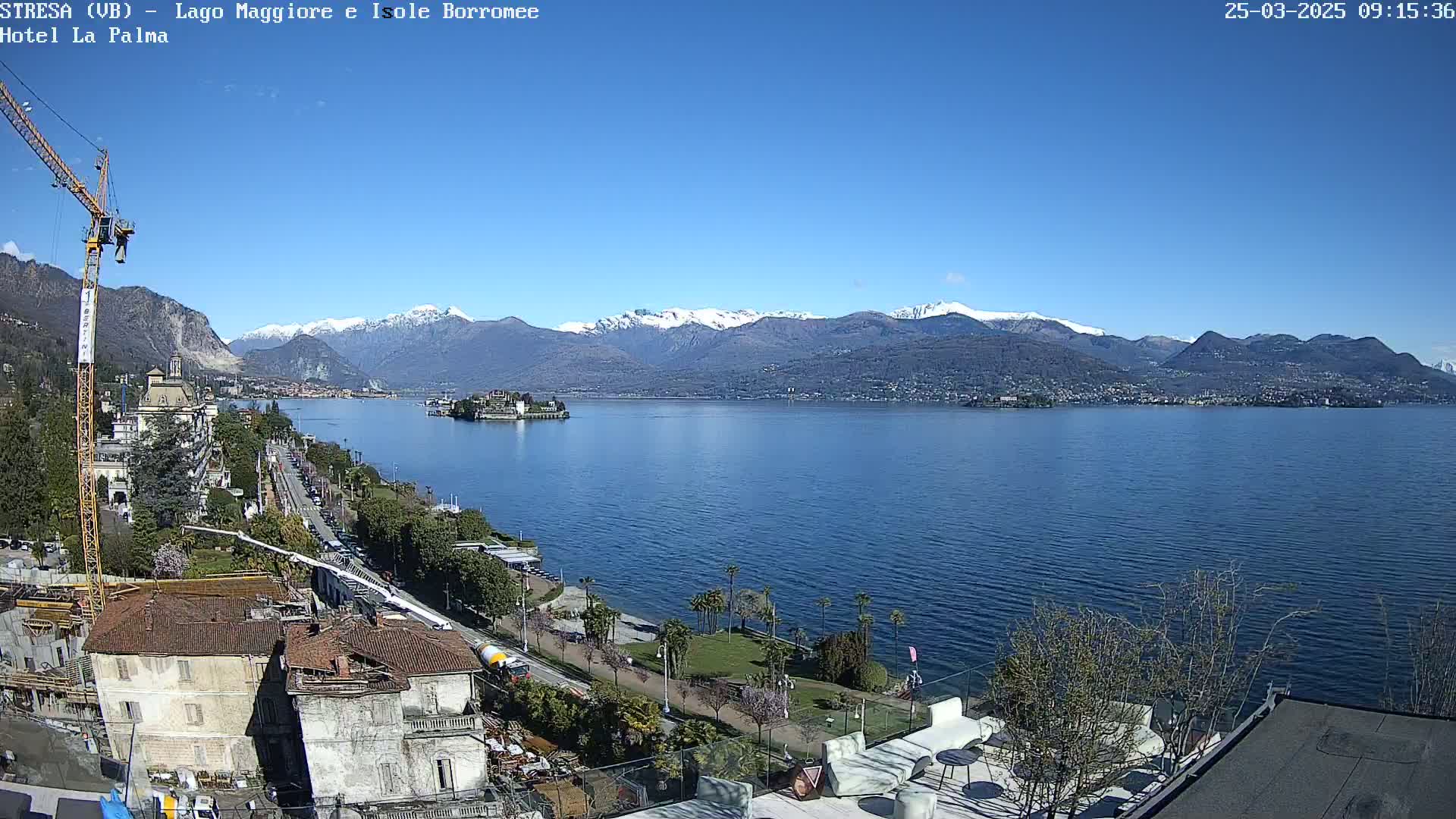 A sunny day overlooking a calm lake with snow-capped mountains in the background, a shoreline with buildings and trees, and a construction crane visible.