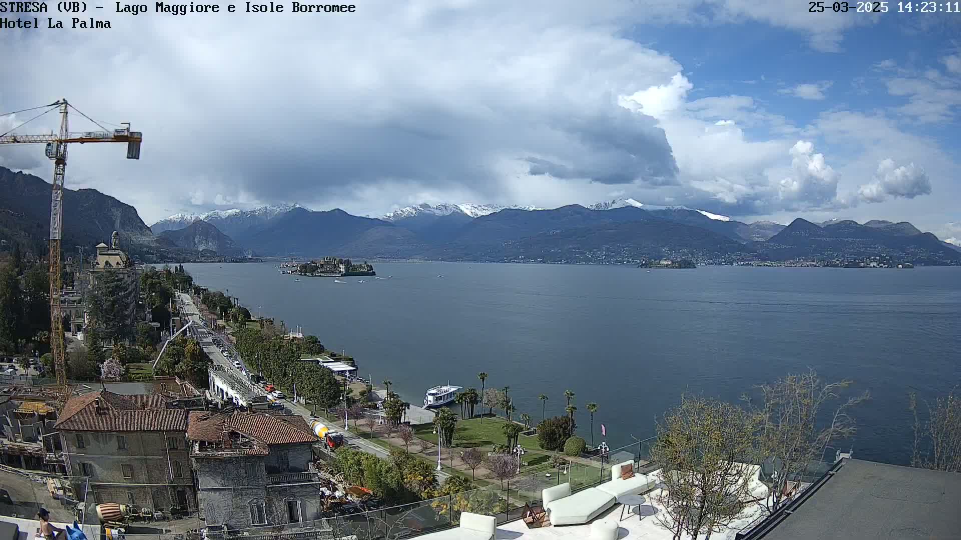 A partly cloudy day reveals a view of a lake with snow-capped mountains in the background, a small island, and buildings along the shoreline.