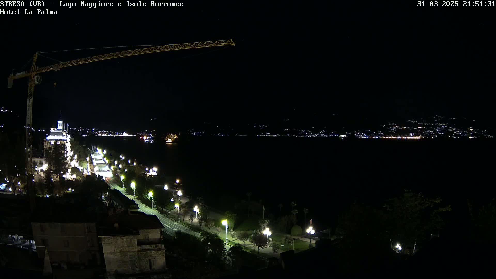 A nighttime, high-angle view of a city along a body of water, illuminated by streetlights, with a construction crane visible in the upper left corner.