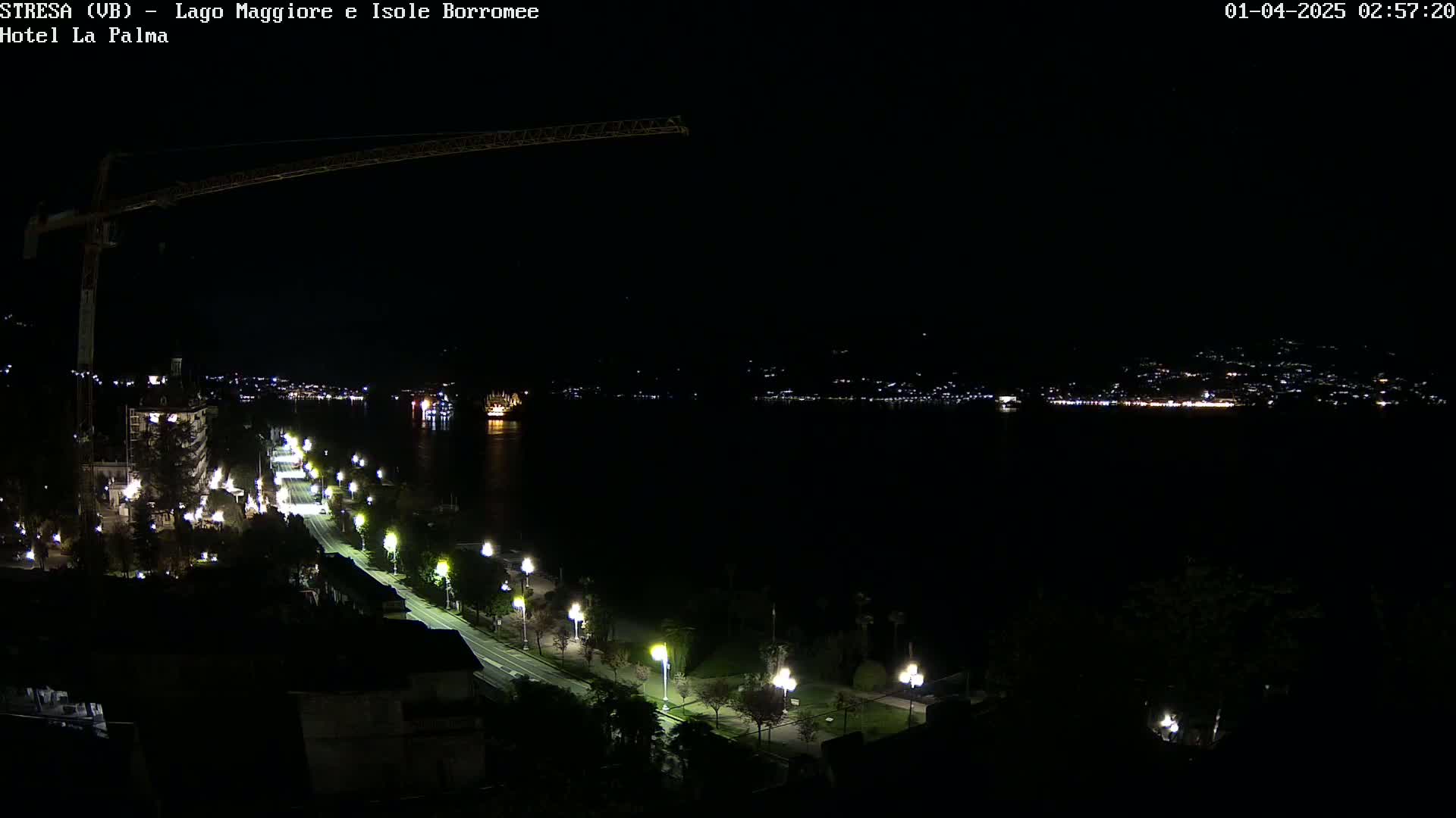 A nighttime view of a city along a dark body of water, illuminated by streetlights and distant city lights, with a construction crane visible in the upper left.