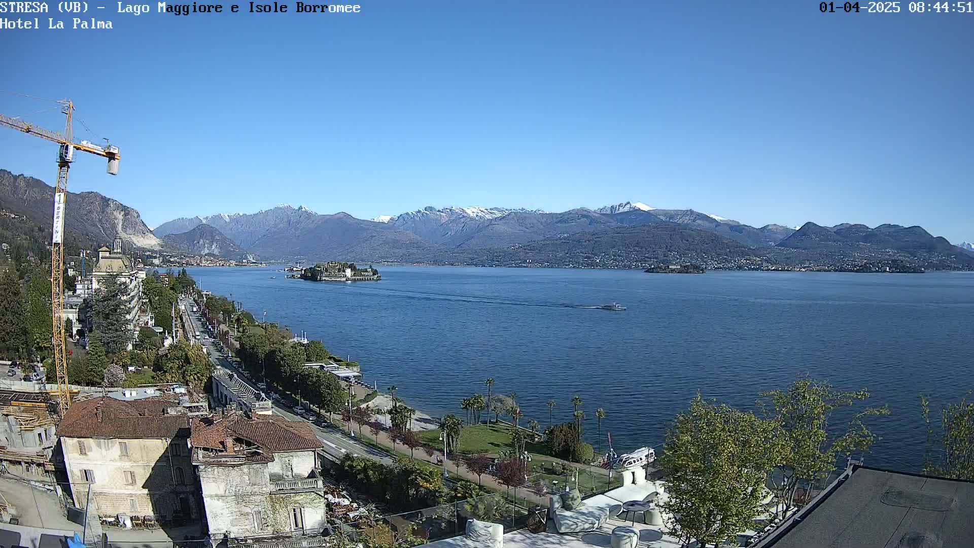 A clear, sunny day overlooks a lake with snow-capped mountains in the background, a small island in the middle of the lake, and buildings and trees along the lakeshore.